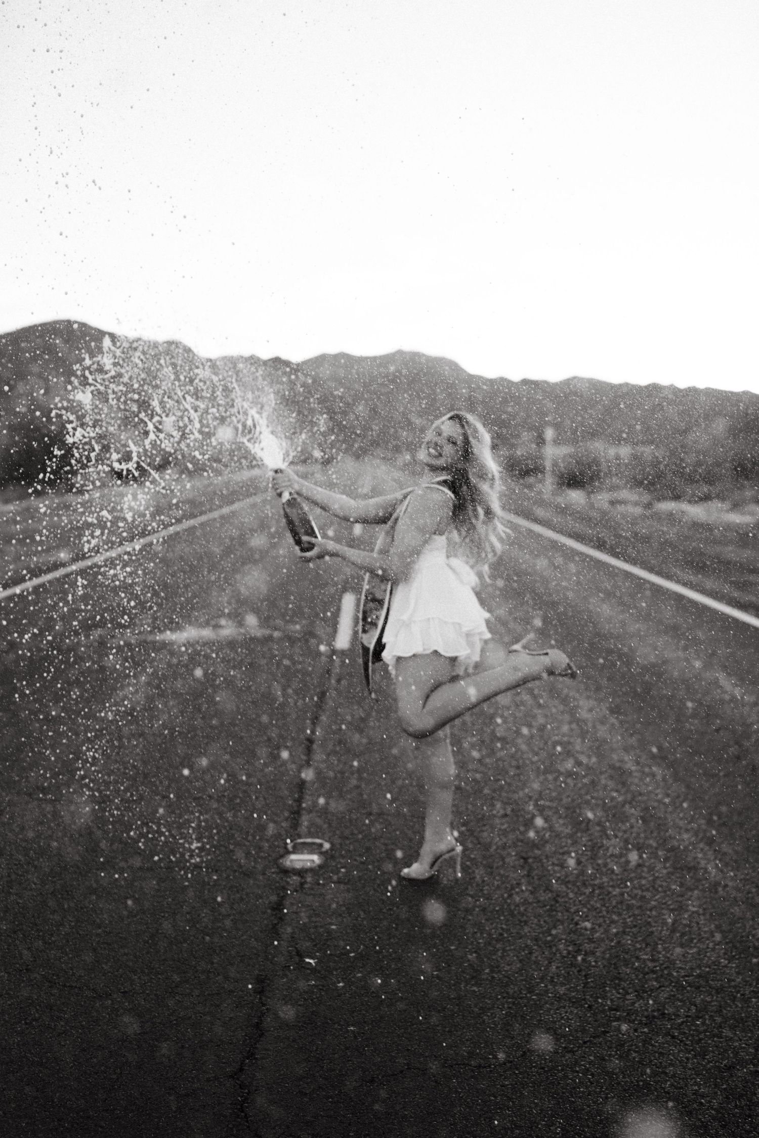 Graduate jumping joyfully while spraying champagne on a desert road in black and white.