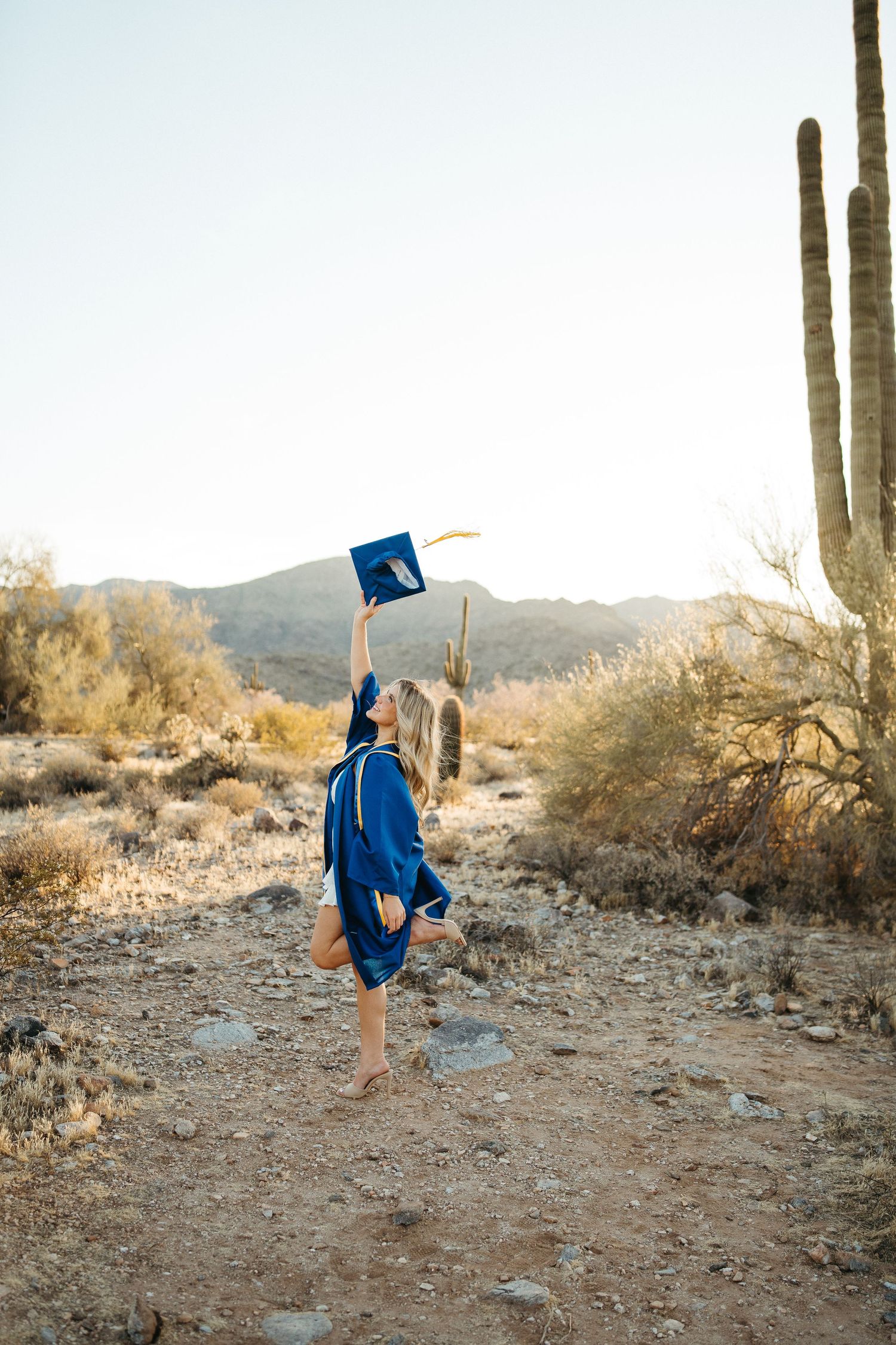 Graduate in blue cap and gown throws graduation cap in desert setting with saguaro cactus.