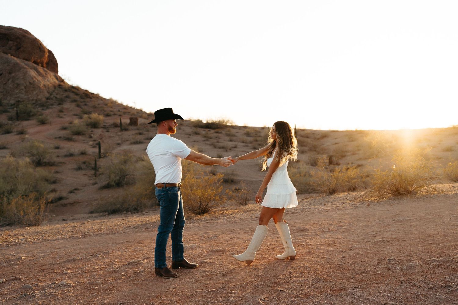 Silhouetted couple strolls through desert terrain as sun sets behind rocky mountain landscape.