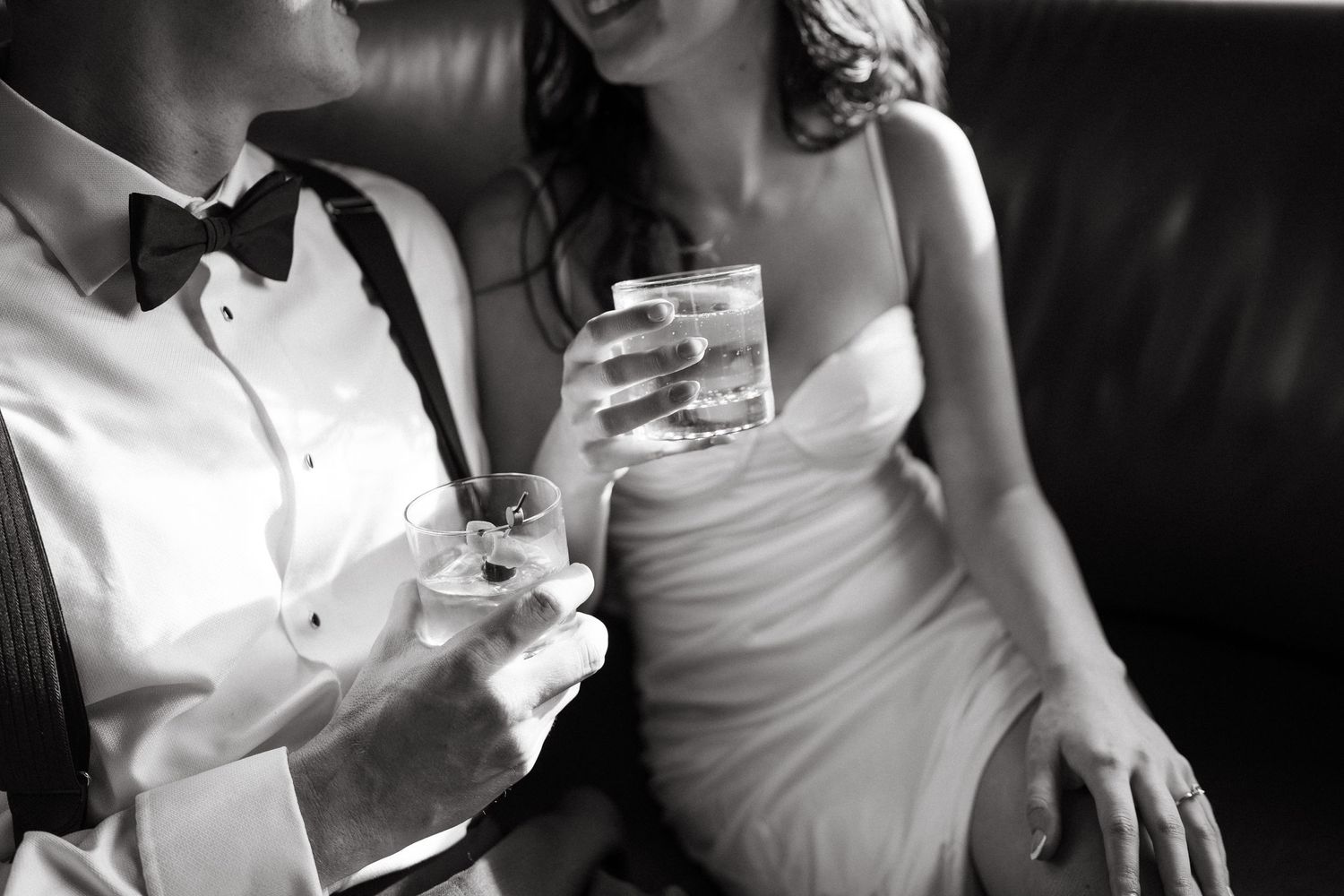 A black and white photo of a couple sharing drinks on leather couch.