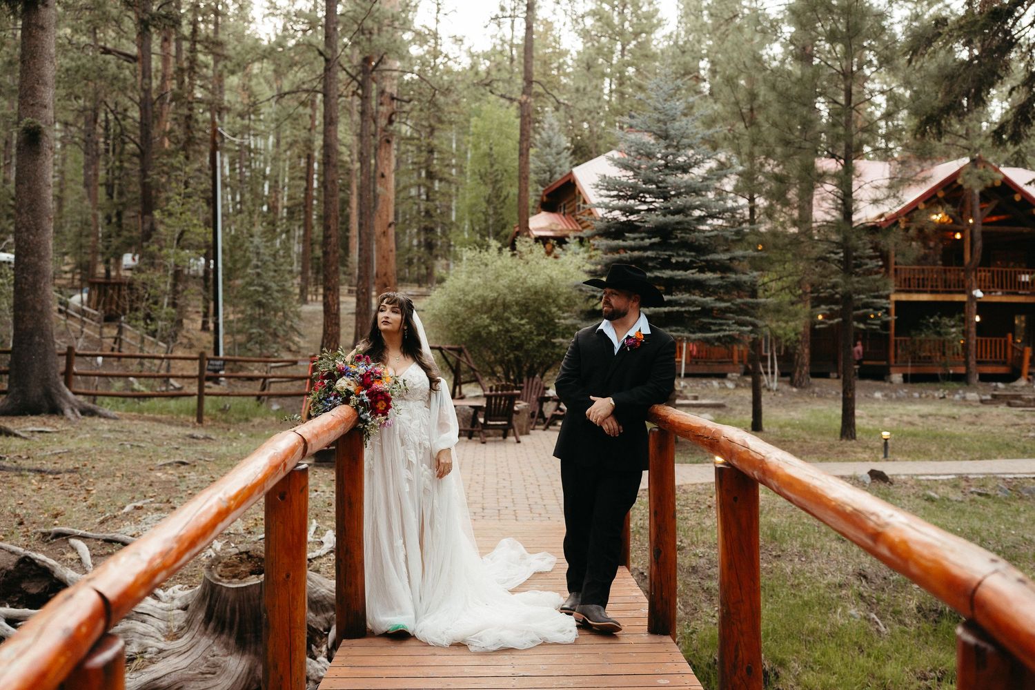 A couple stands on a rustic wooden bridge surrounded by tall pine trees in a forest wedding setting.