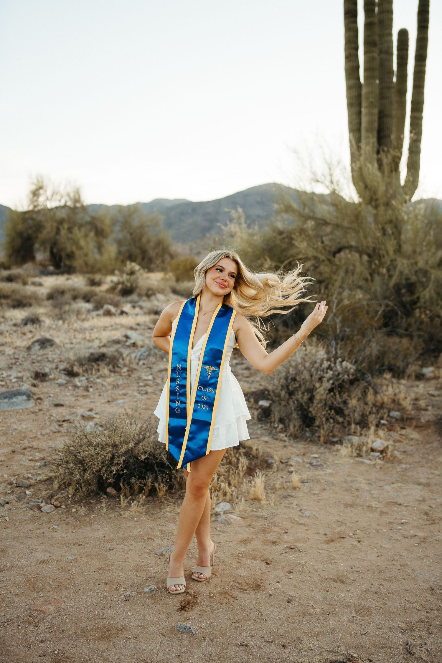 Graduate celebrates in a white dress and blue stole while posing in a desert landscape with saguaro cactus at sunset.
