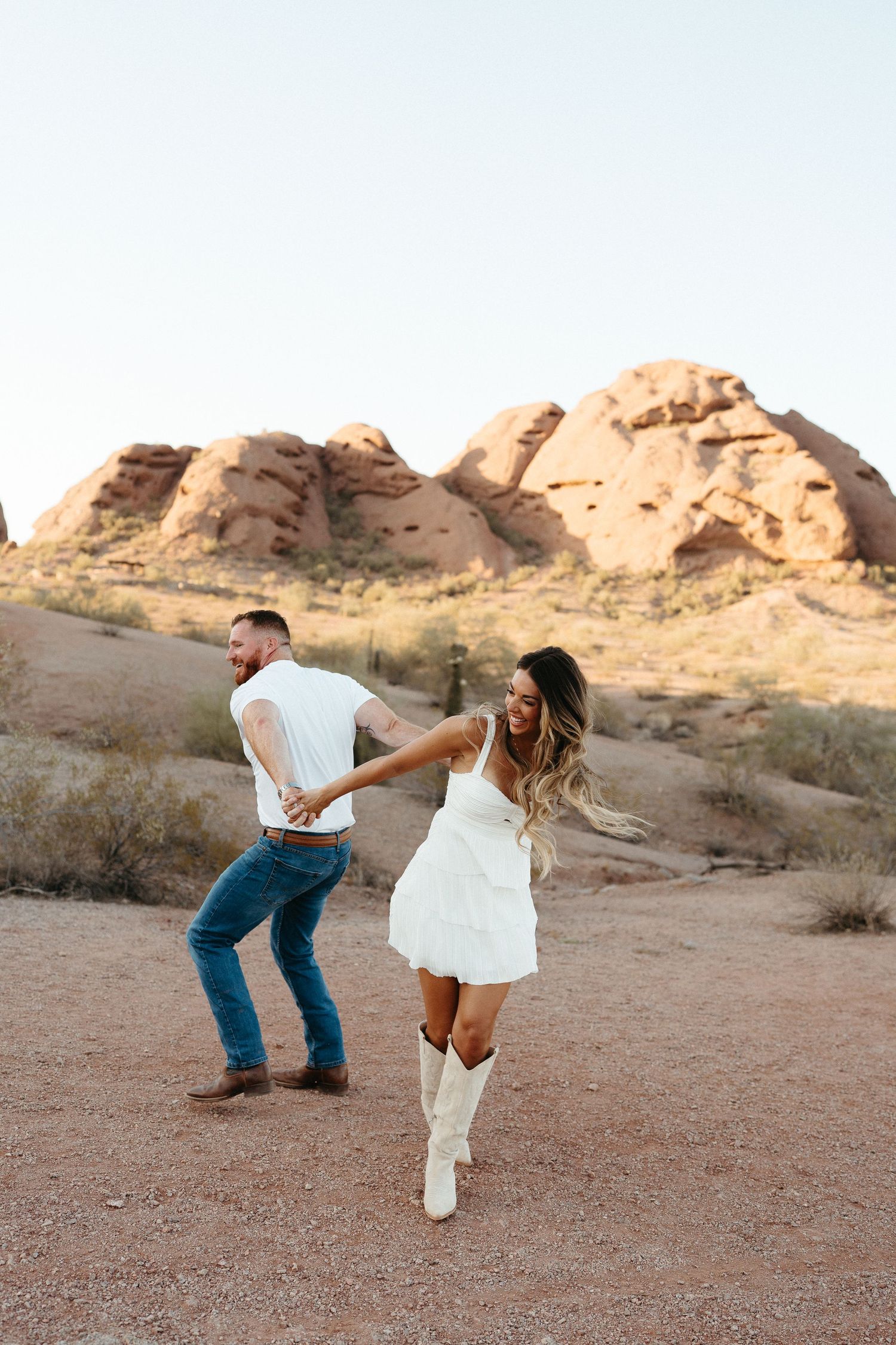 A couple dances playfully in white western attire against scenic desert rock formations at sunset.