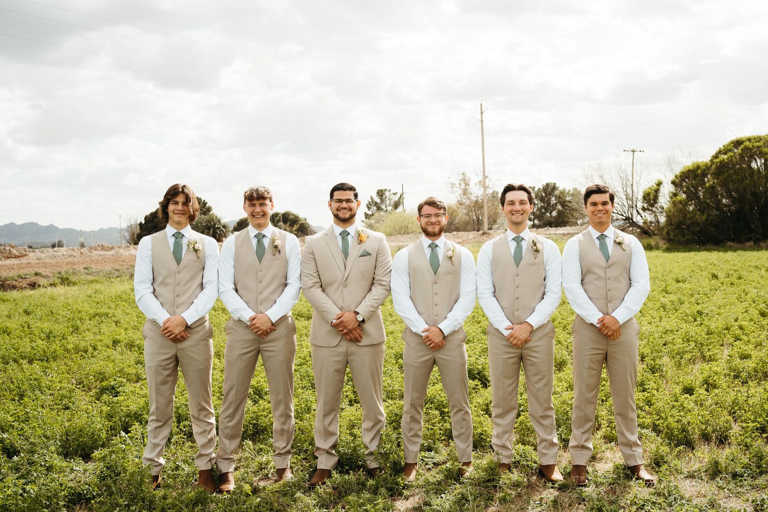 Groomsmen in light beige suits pose together on a grassy hillside under cloudy skies.
