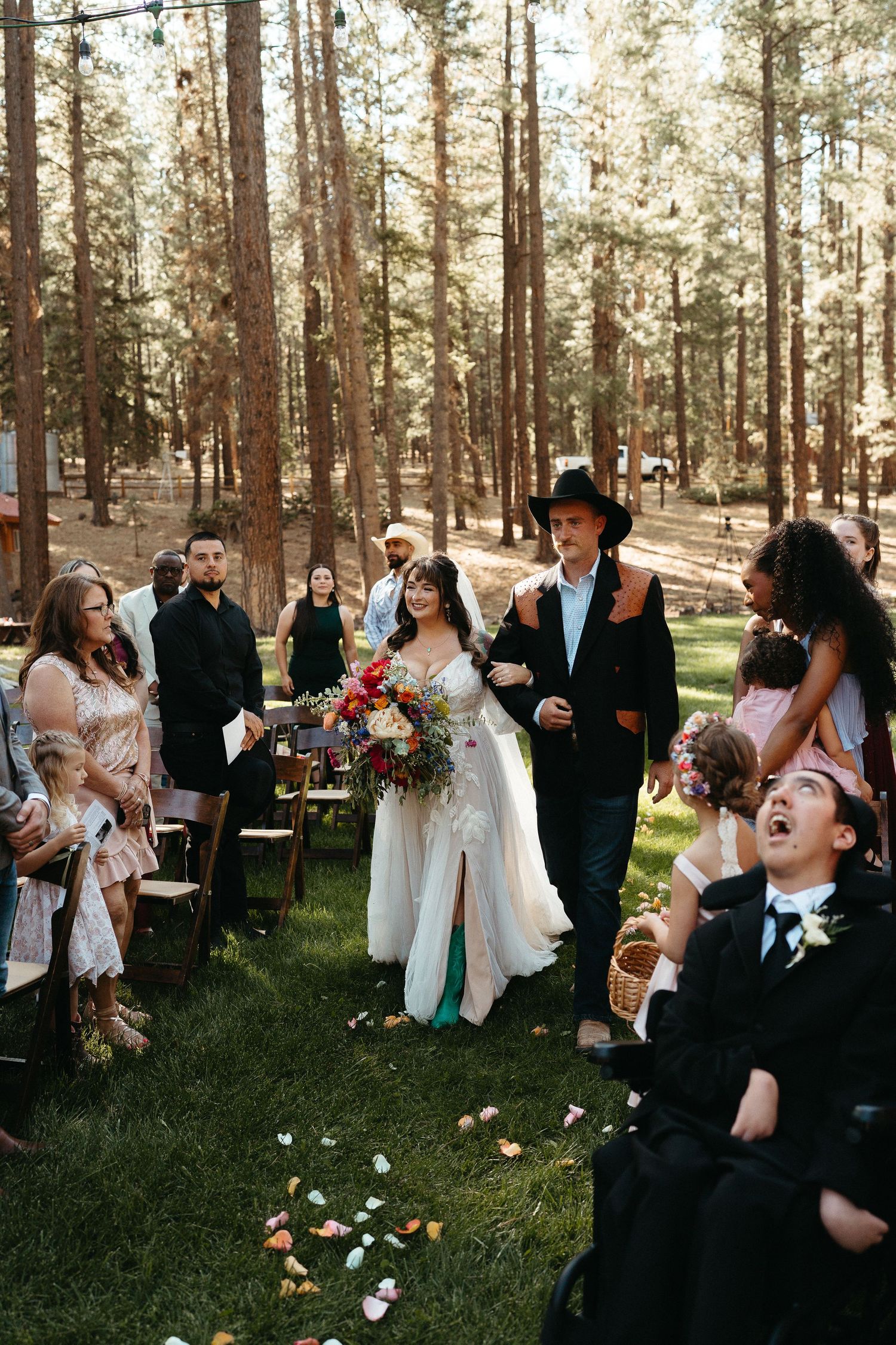 Wedding party celebrates in a sunlit forest setting with flower petals being tossed in the air.