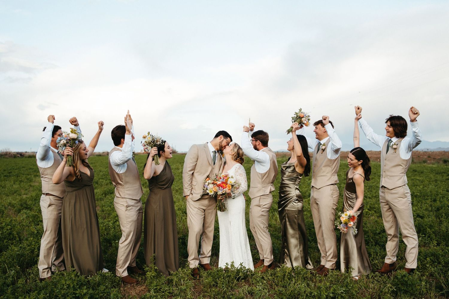 Wedding party in beige attire posing with raised arms against cloudy sky.