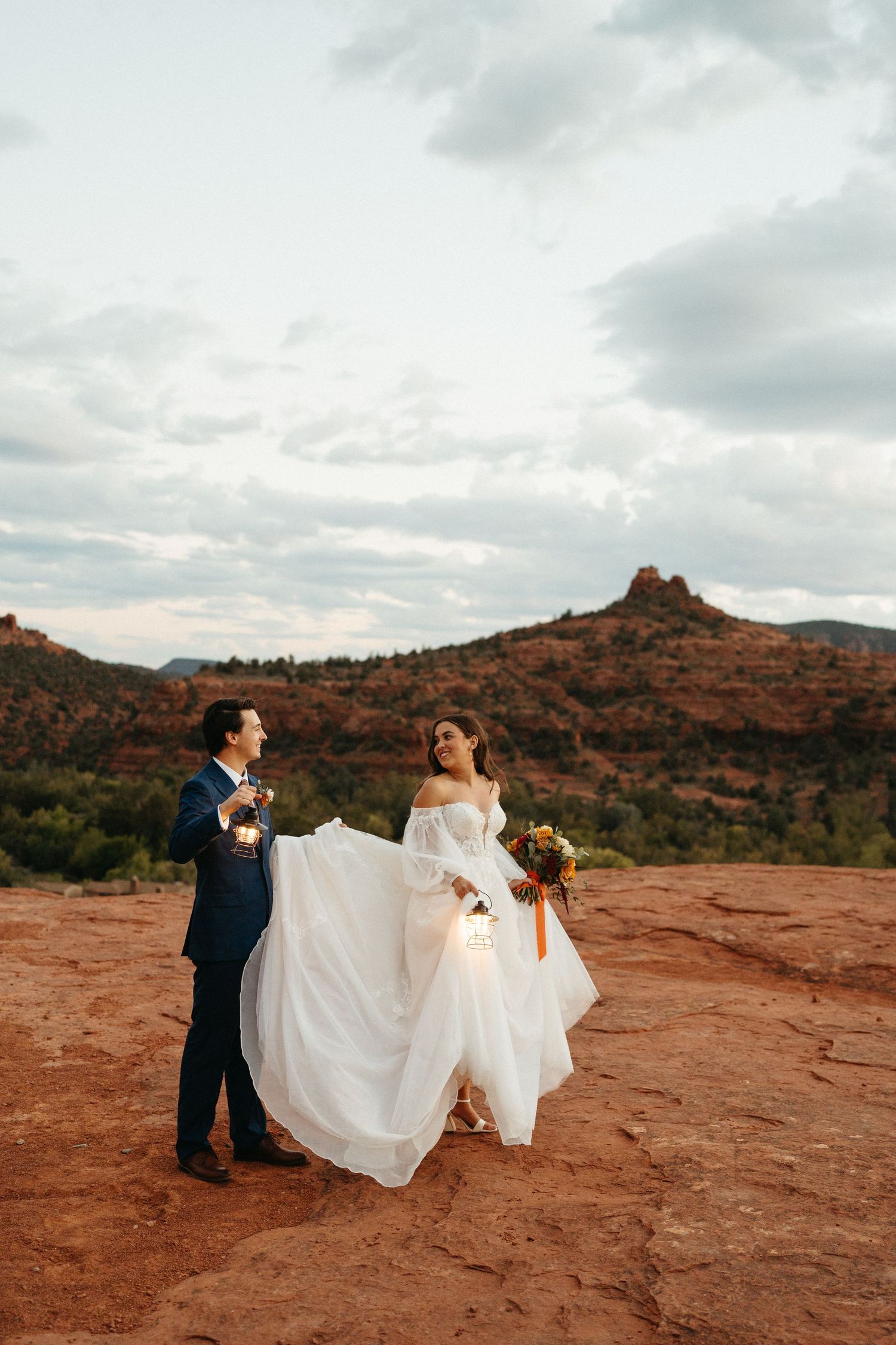 A couple in wedding attire poses on red rock formations at sunset in Sedona, Arizona with mountains in the background.