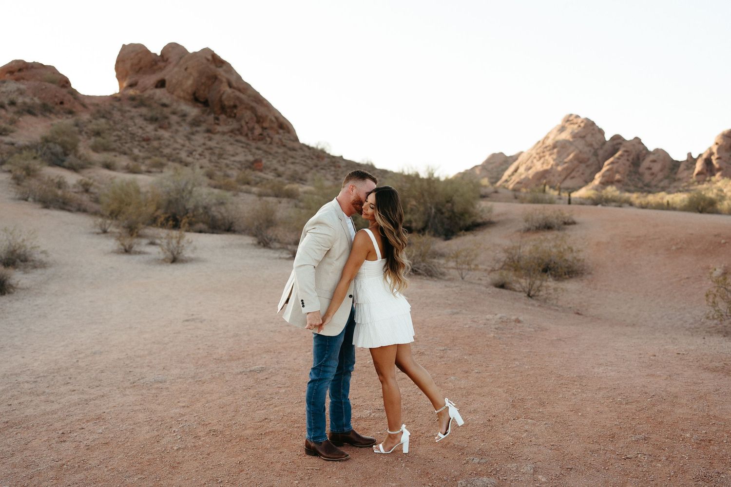 Two figures embrace and kiss in a romantic moment against the backdrop of red desert rock formations at sunset.