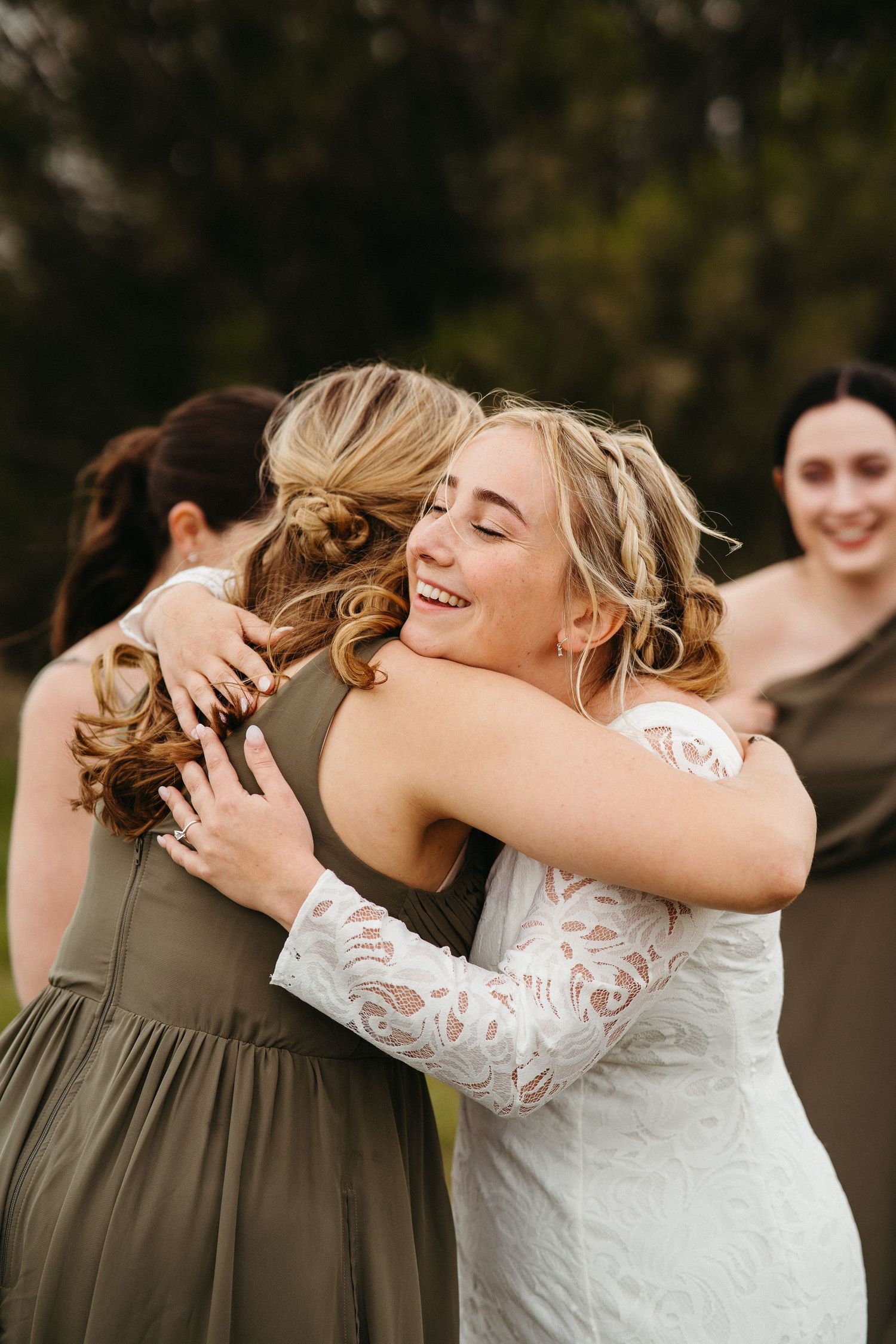 An emotional embrace shared between a bride in white lace and bridesmaid in olive green.
