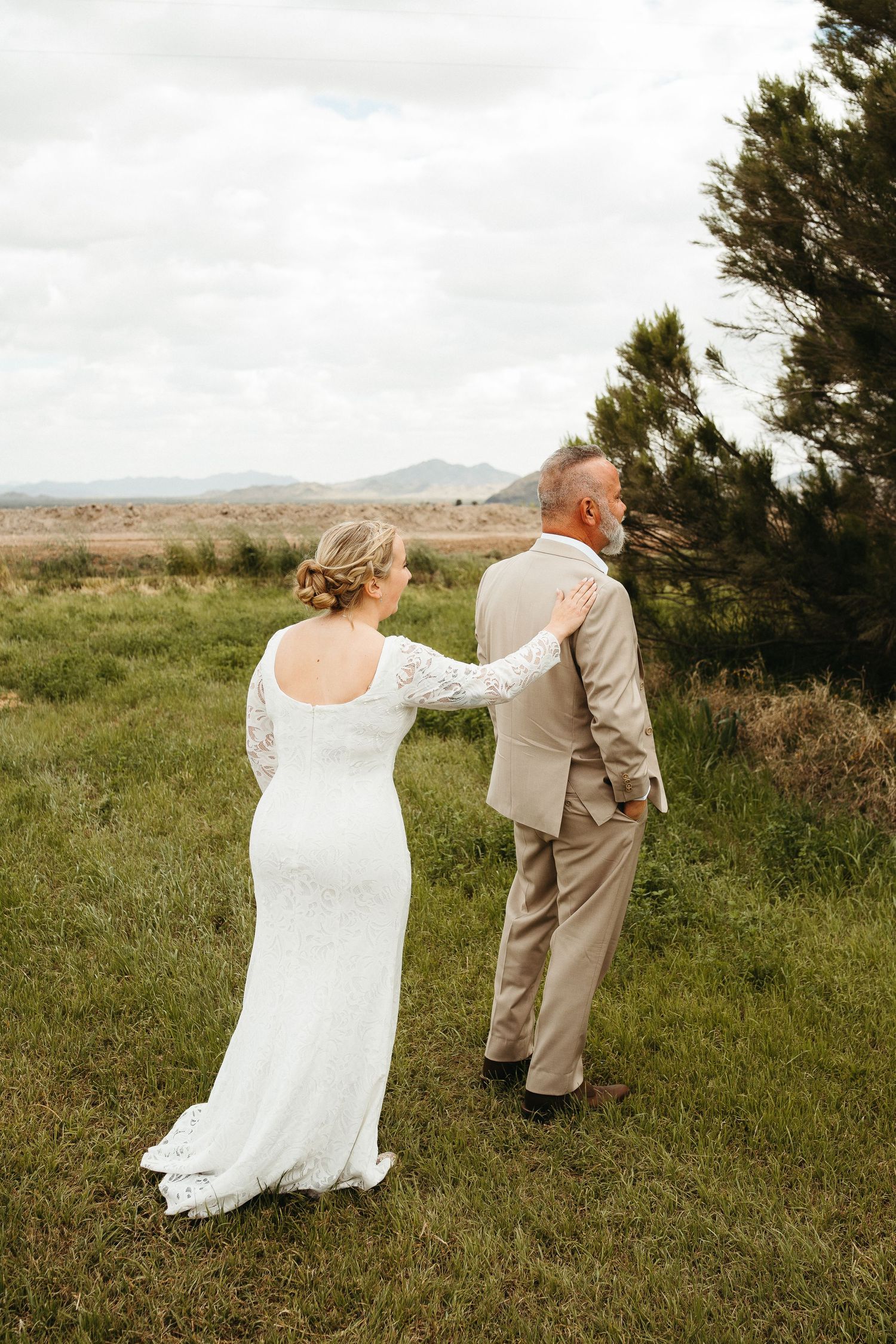 A couple embraces while looking at mountain views during their wedding photoshoot in a grassy field.