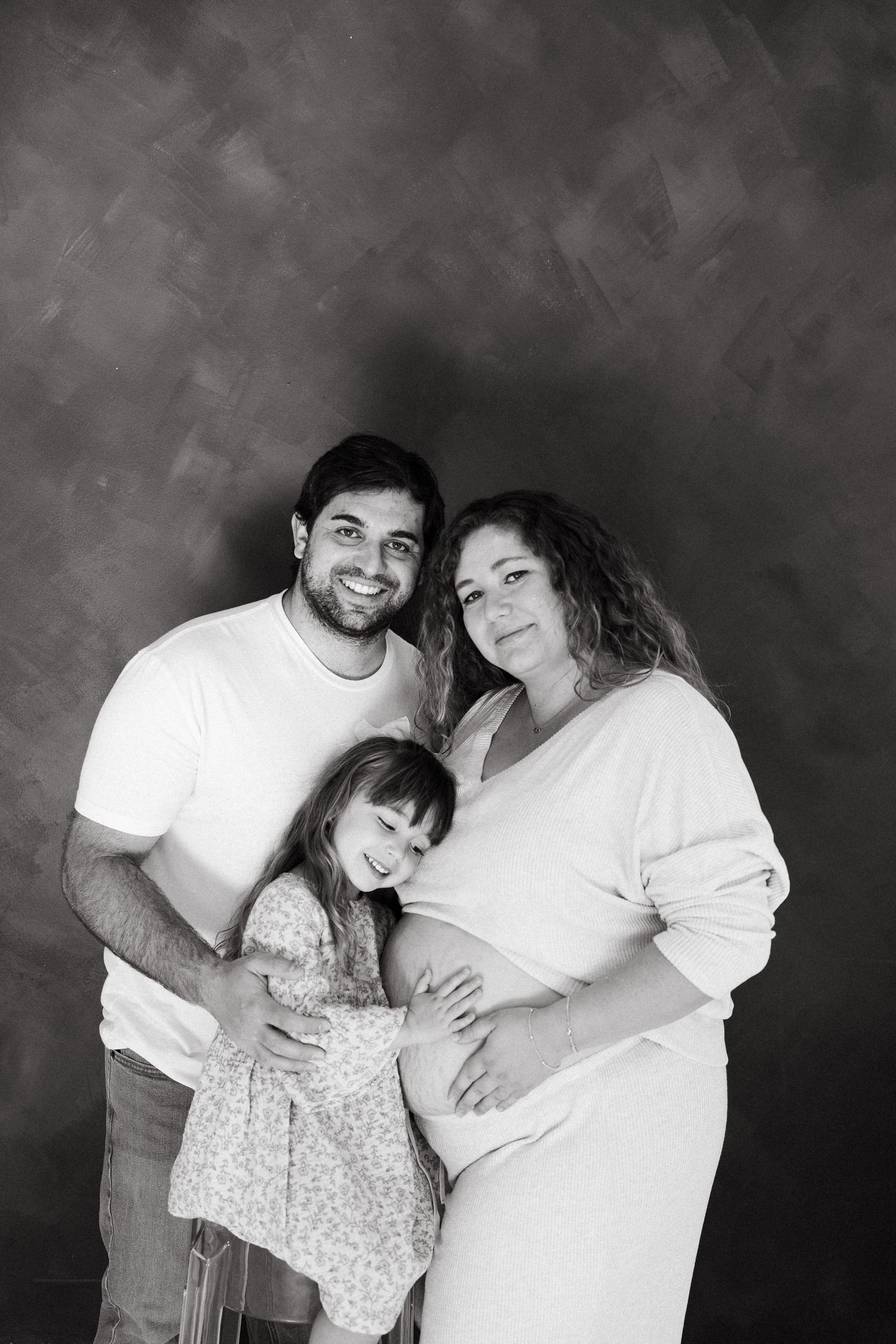 A black and white portrait showing a family of three posing together, with a small child in a floral dress.