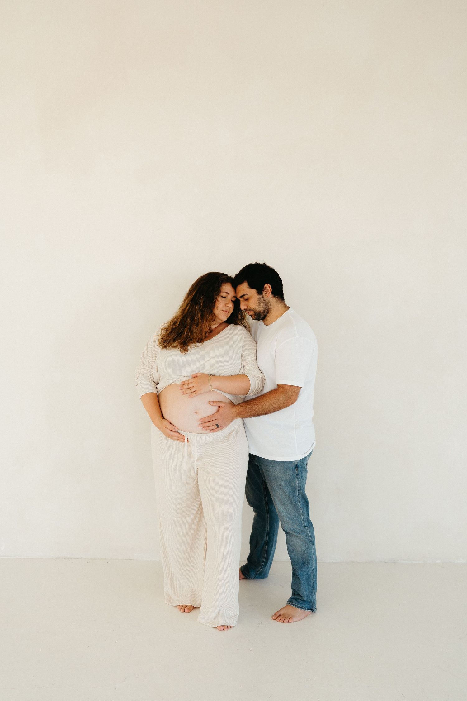 A couple in casual attire shares tender embrace against plain light-colored wall in minimalist studio setting.