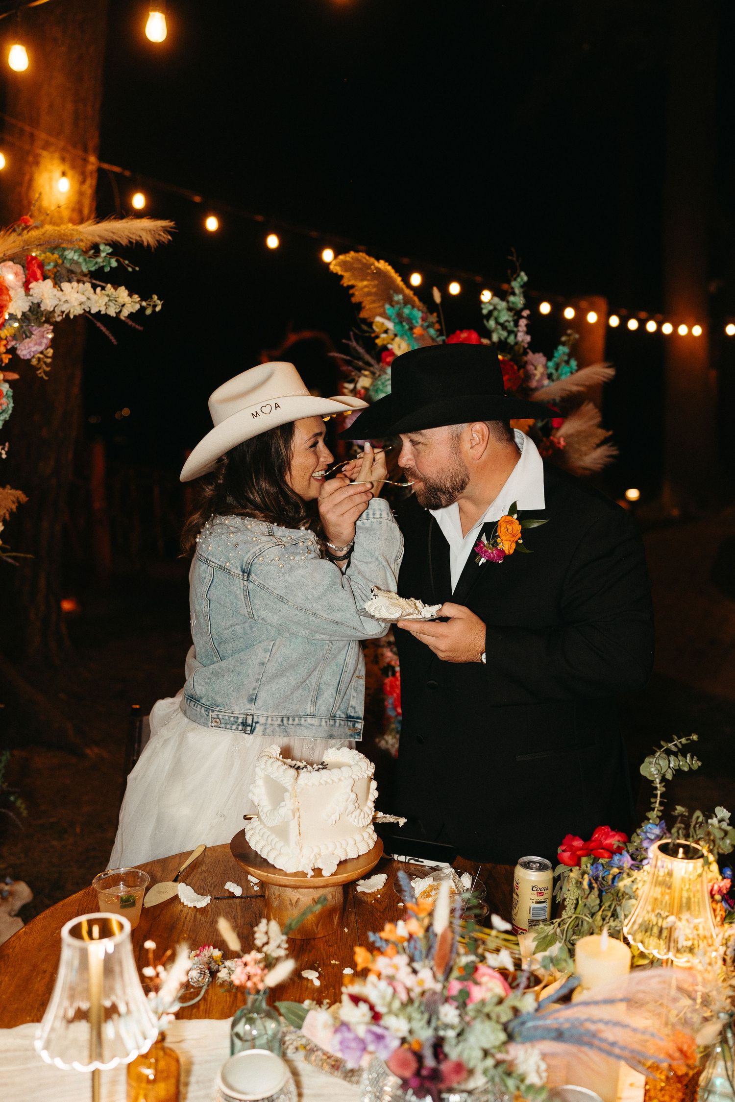 Couple in western attire share cake at rustic outdoor wedding reception with string lights and floral decorations.