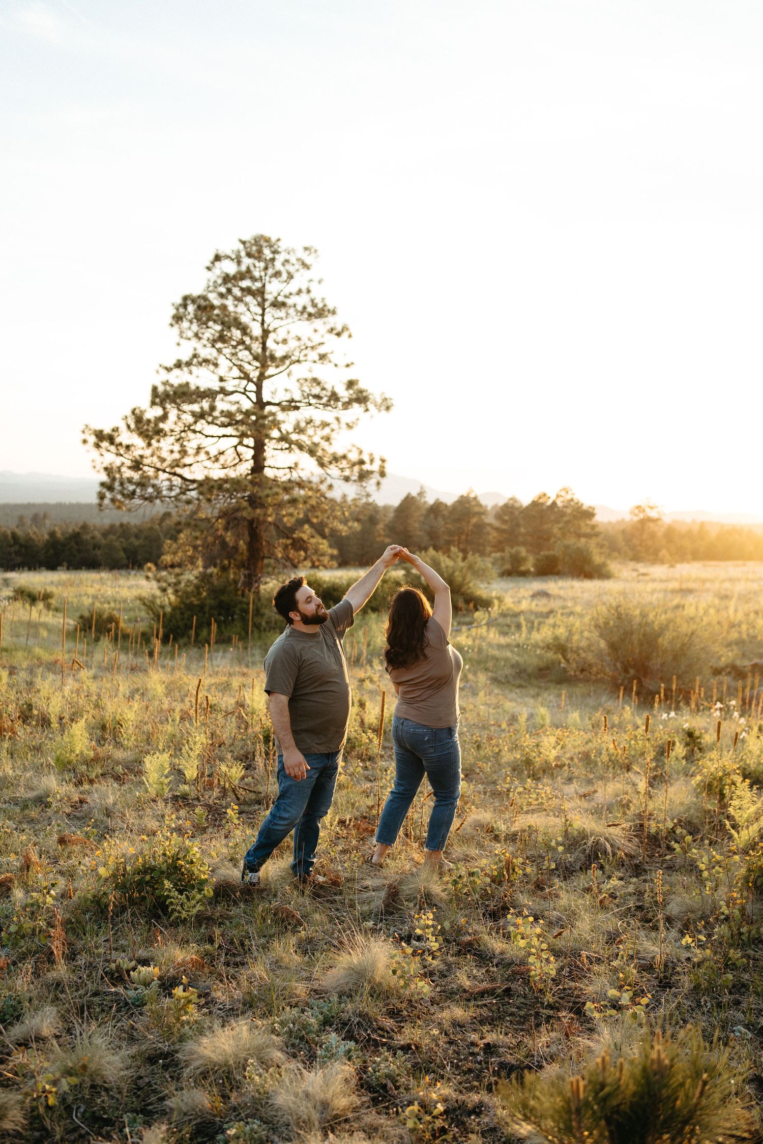 A couple dancing together in a golden field at sunset with a large pine tree in the background.