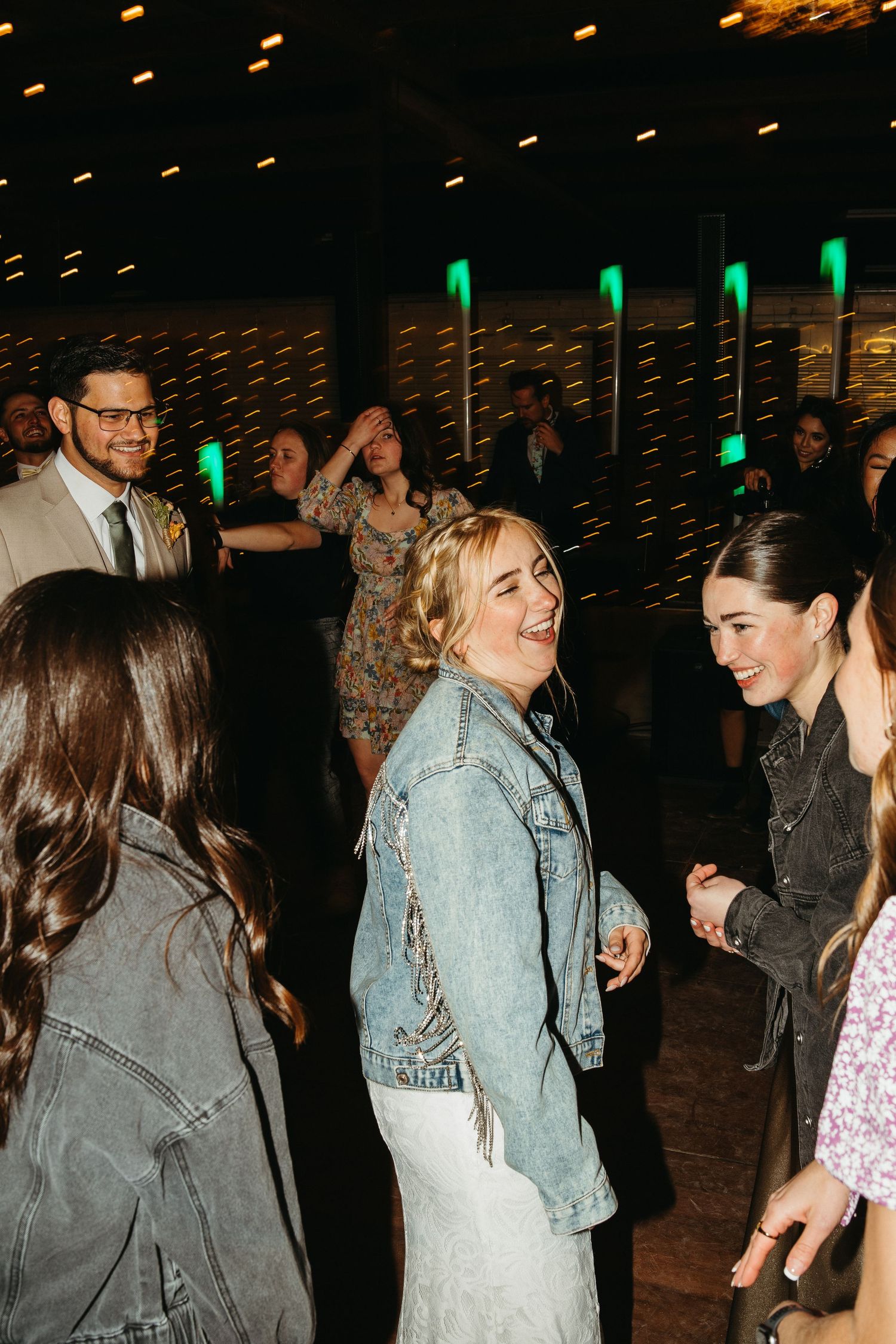 Wedding guests dancing and laughing together under moody lighting at an indoor reception venue.