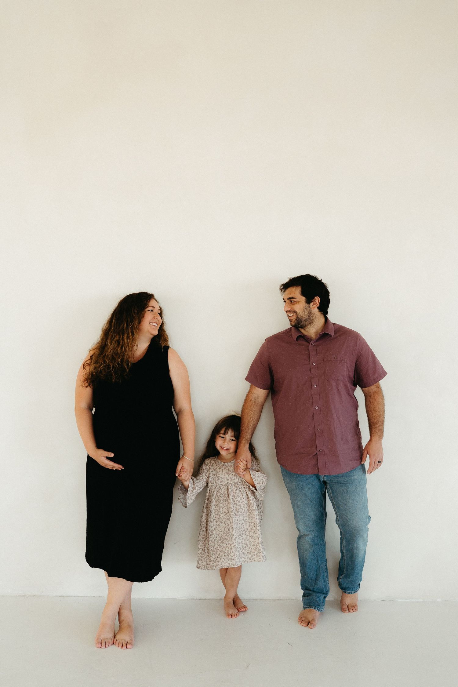 A family stands relaxed against a white wall, barefoot and smiling at each other while posing for a photo.