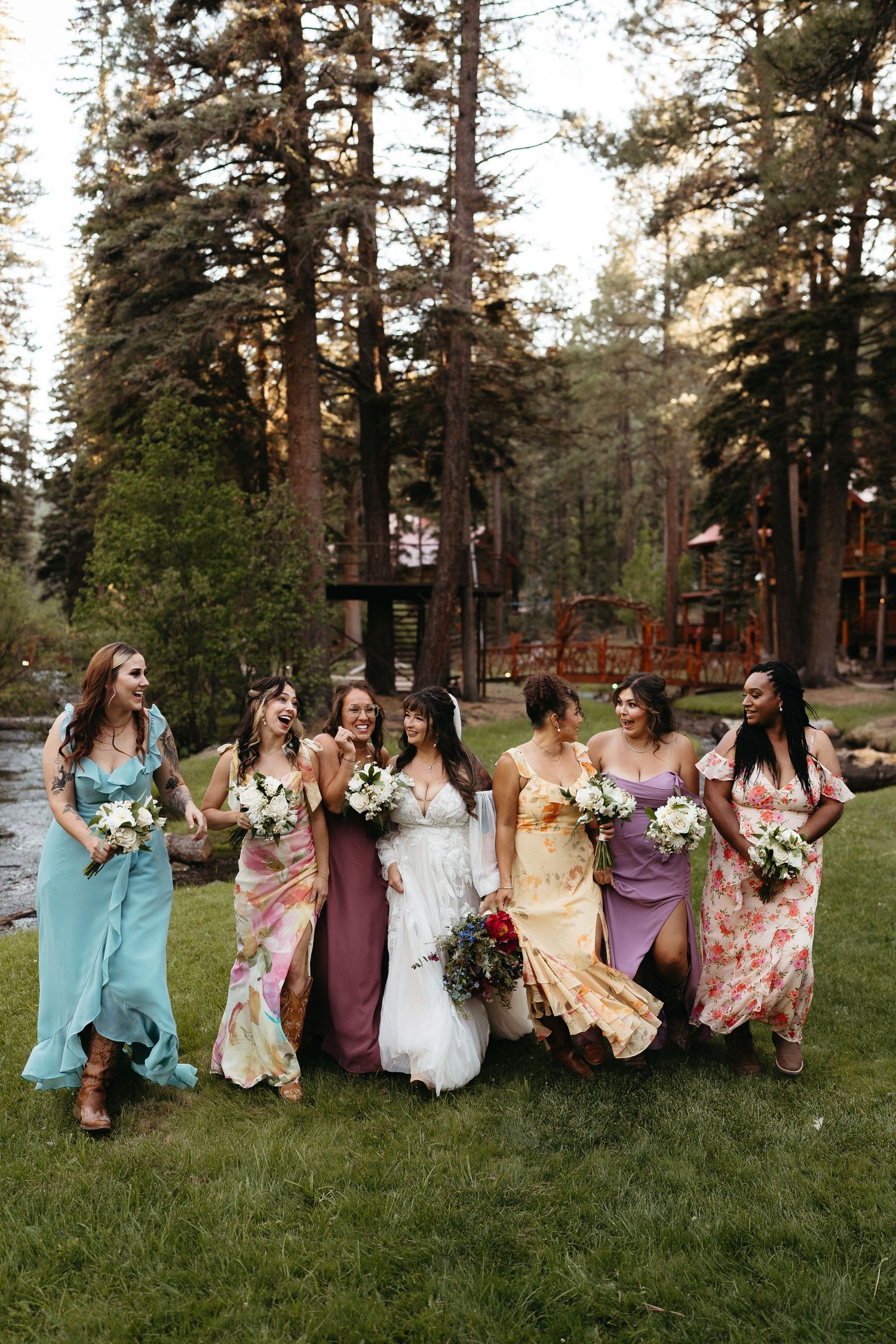 Wedding party members sit casually on grass in coordinated purple and floral dresses during outdoor photo session.