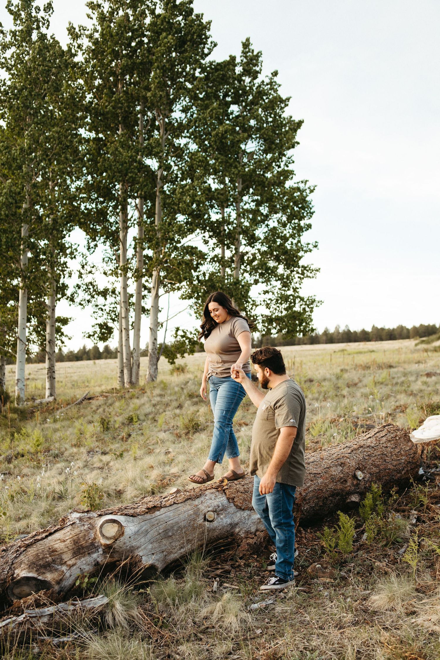 Two people standing on a fallen log amid aspen trees on a grassy hillside.