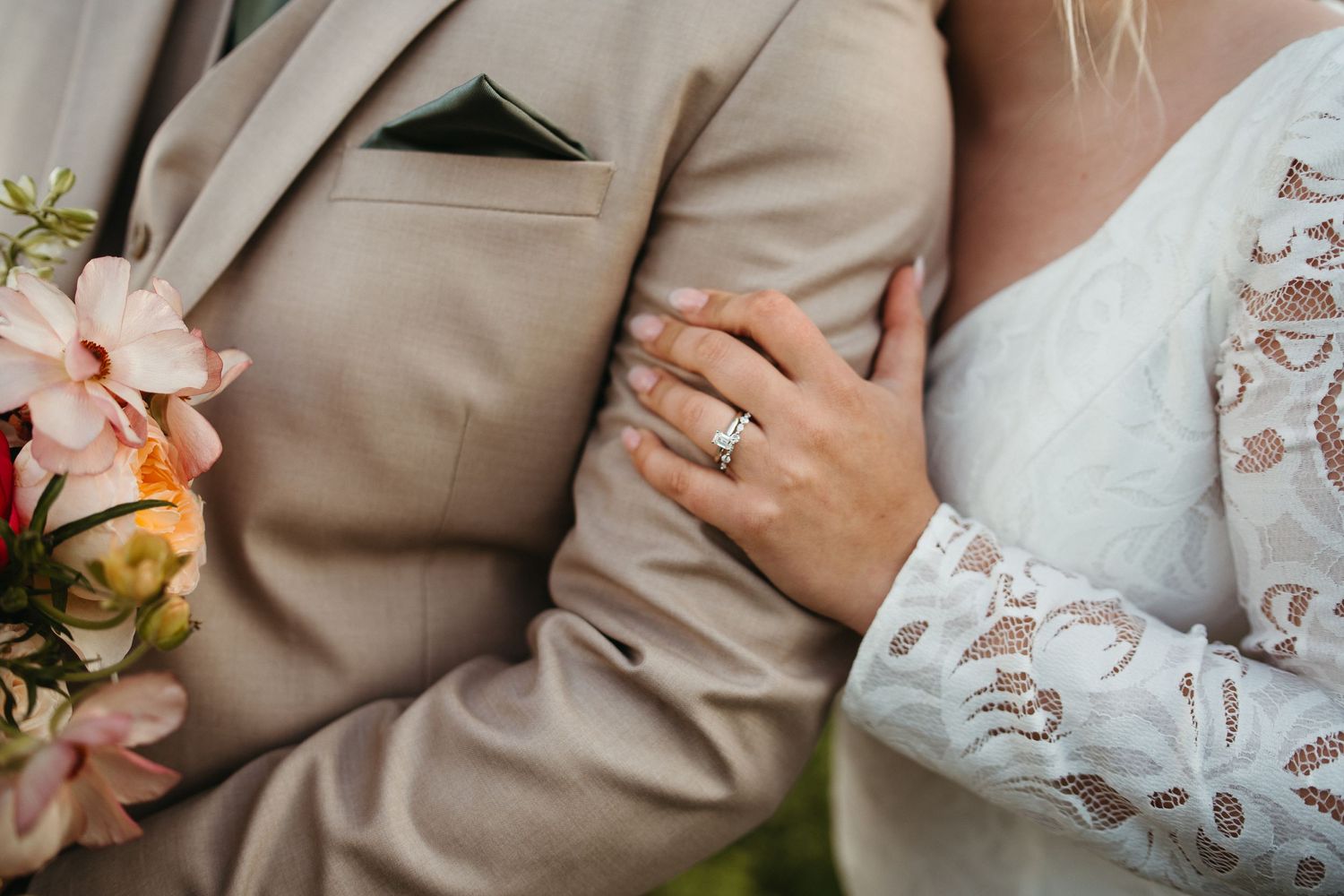 Close-up of a wedding ring and lace sleeve details against a tan suit jacket.