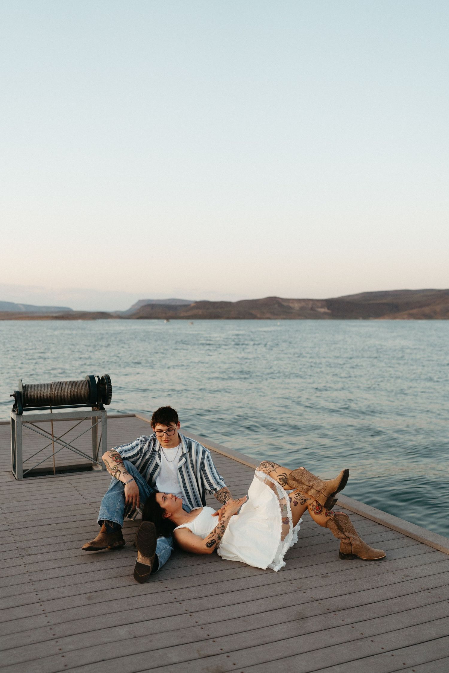 Relaxed scene on lakeside dock with person sitting next to dog and luggage during sunset over calm waters.