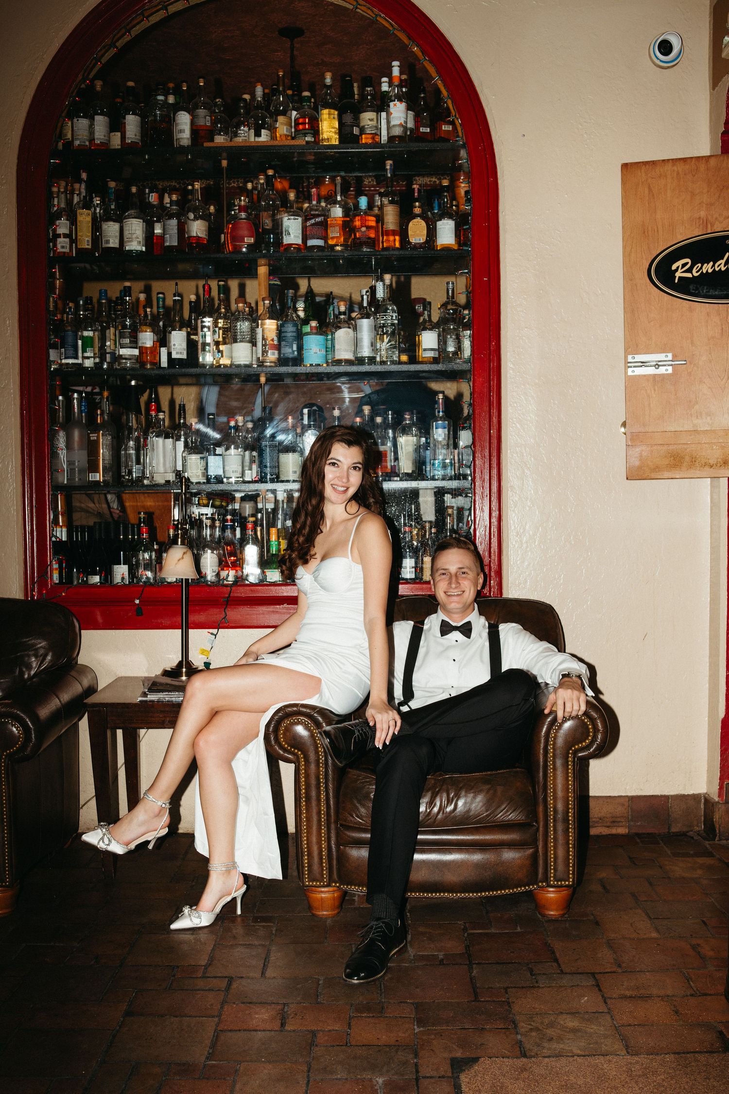 Elegant couple posed in formal attire against vintage bar with extensive liquor collection.