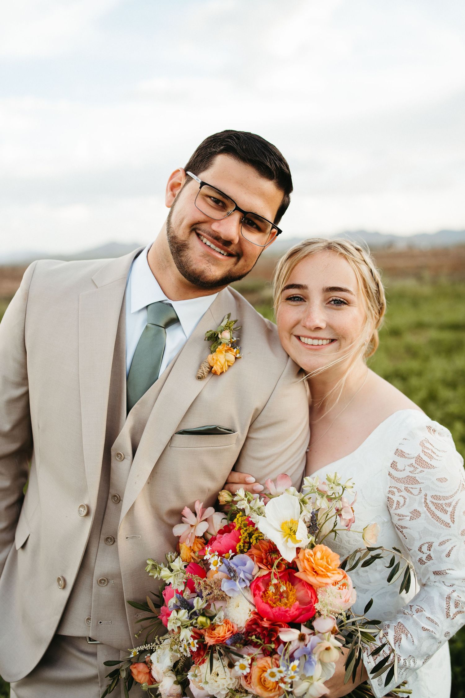 A couple in wedding attire poses outdoors with a vibrant floral bouquet featuring orange and pink blooms.