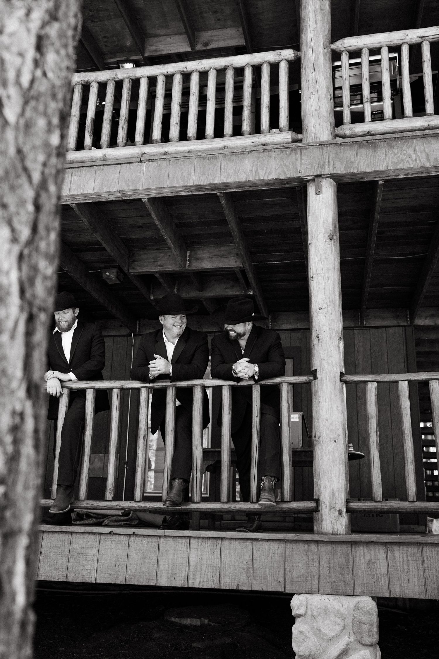 Three figures in dark suits and hats stand on a rustic wooden porch railing in black and white photography.