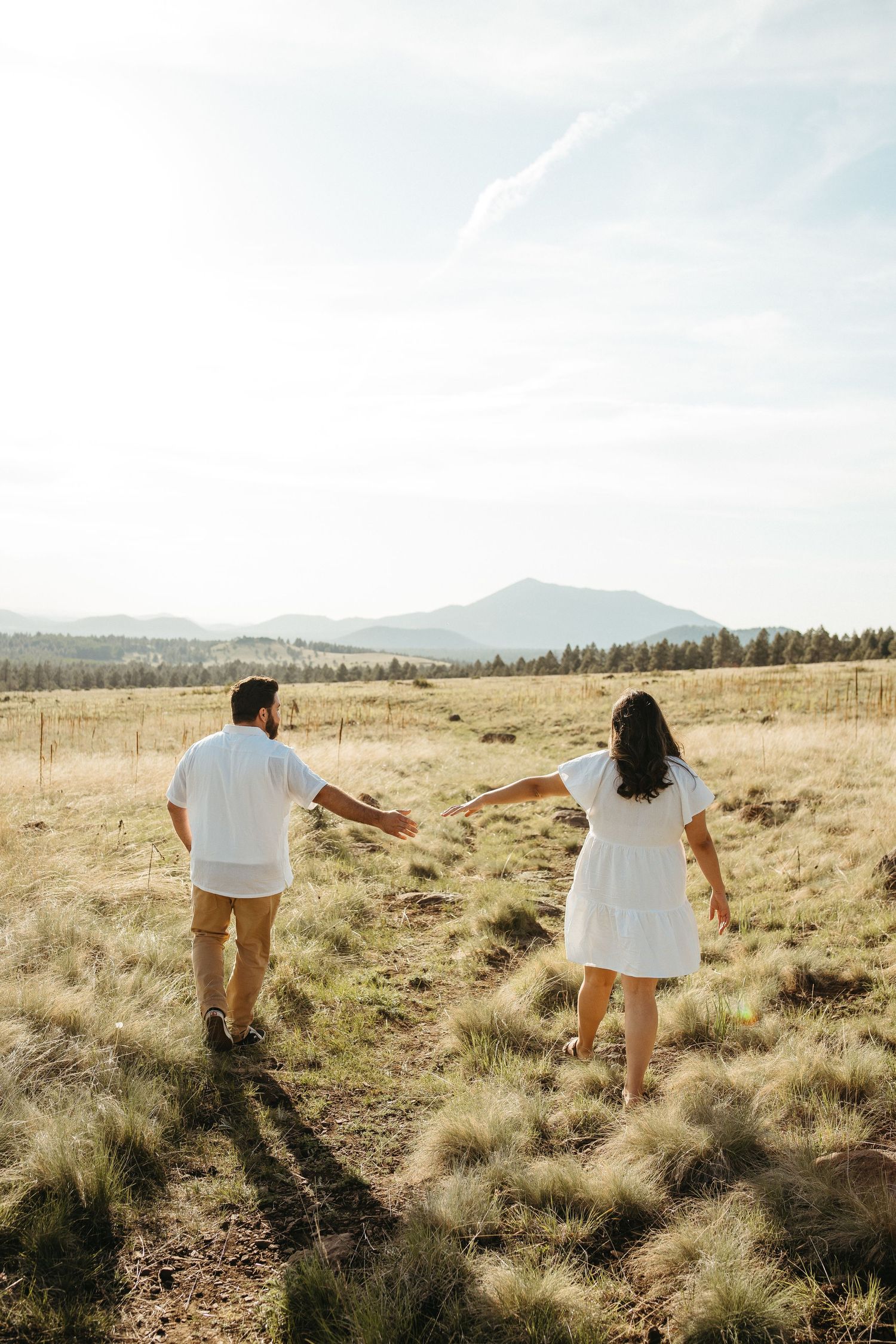 Couple holding hands while walking through golden meadow with mountain view.