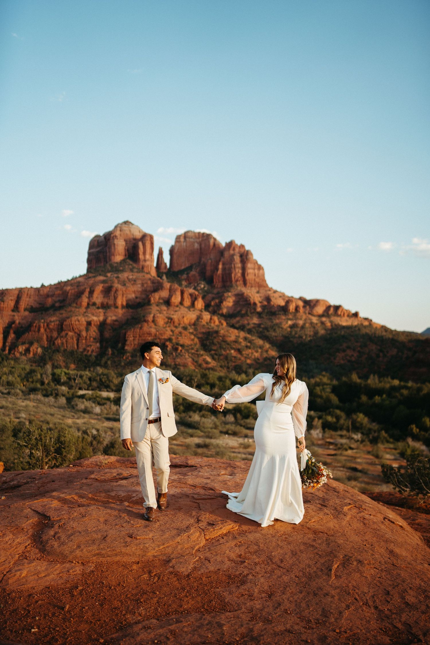 Couple holding hands on red rocks with Cathedral Rock formation in Sedona background.