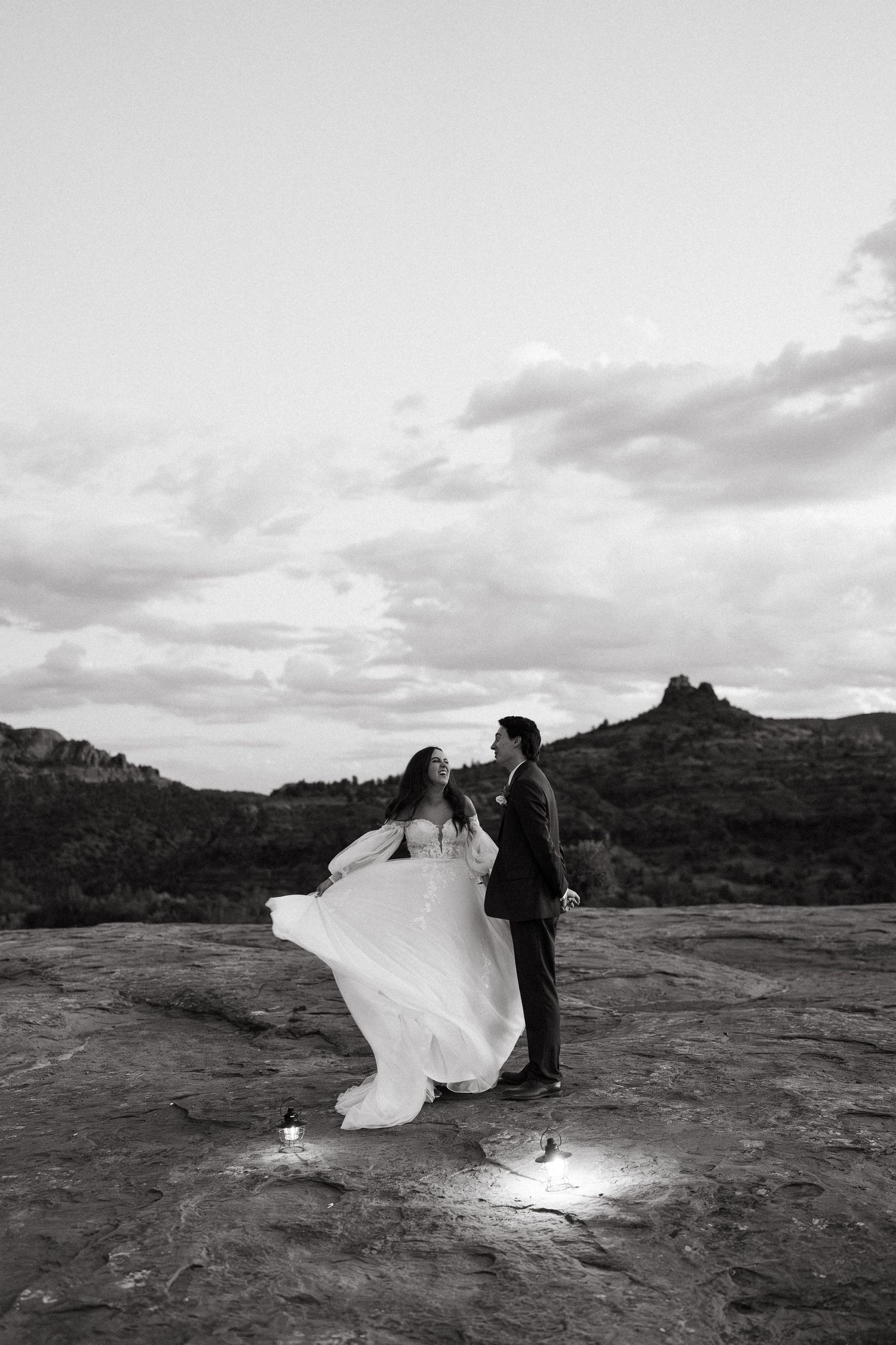 A couple in formal attire poses on red rock formations in Sedona at sunset as the bride's dress flows in the wind.