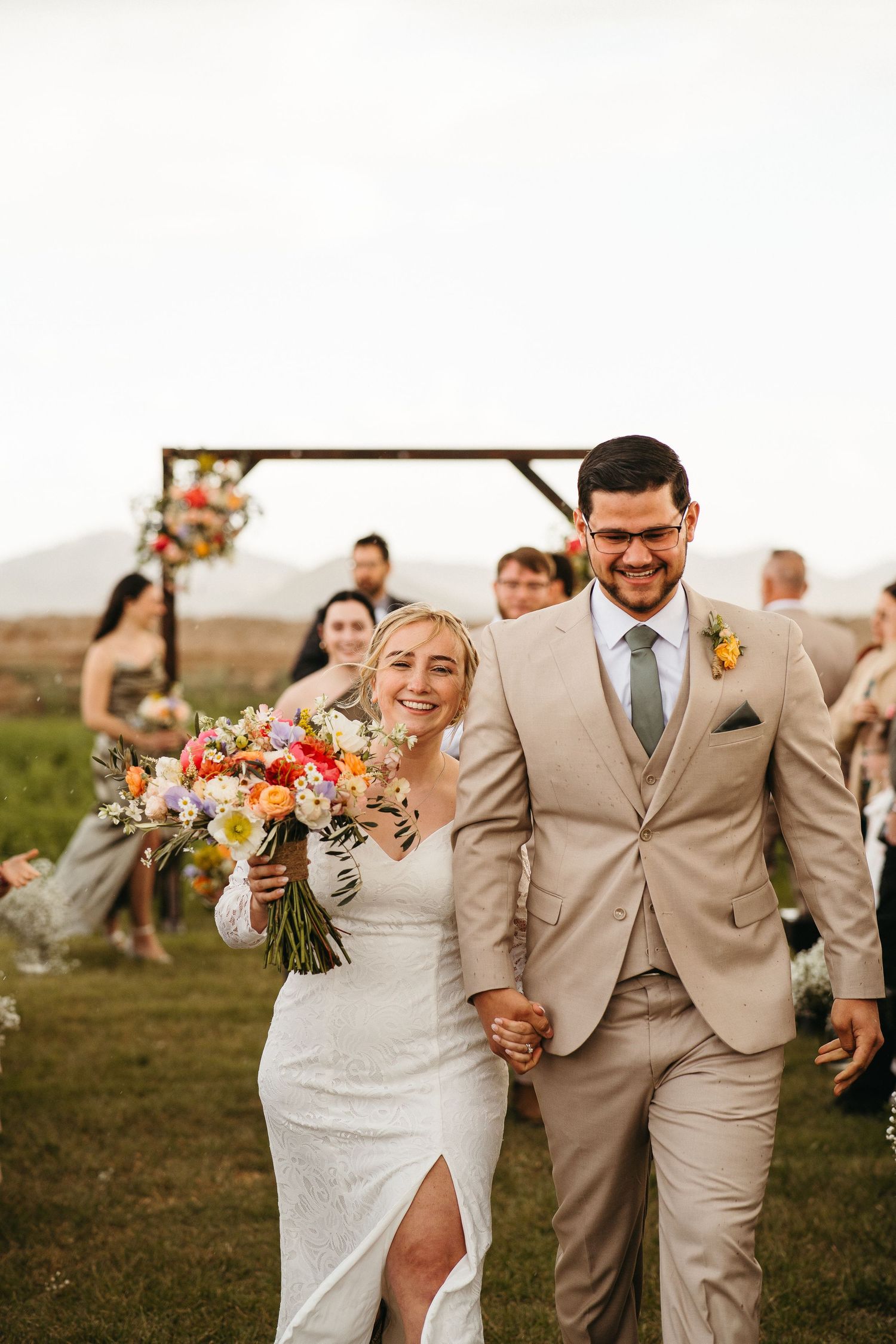 Newlyweds walking down aisle after outdoor ceremony with bridal party in beige attire.
