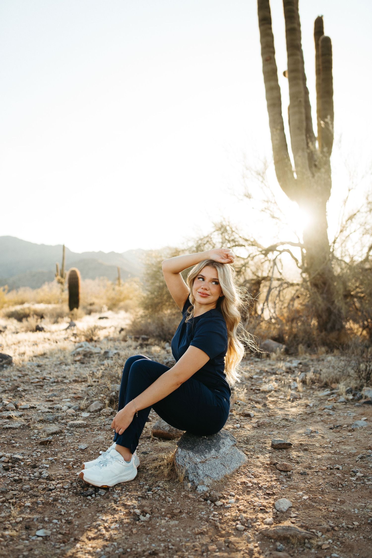 Figure sitting on a rock in the desert with saguaro cacti and mountains in the background.