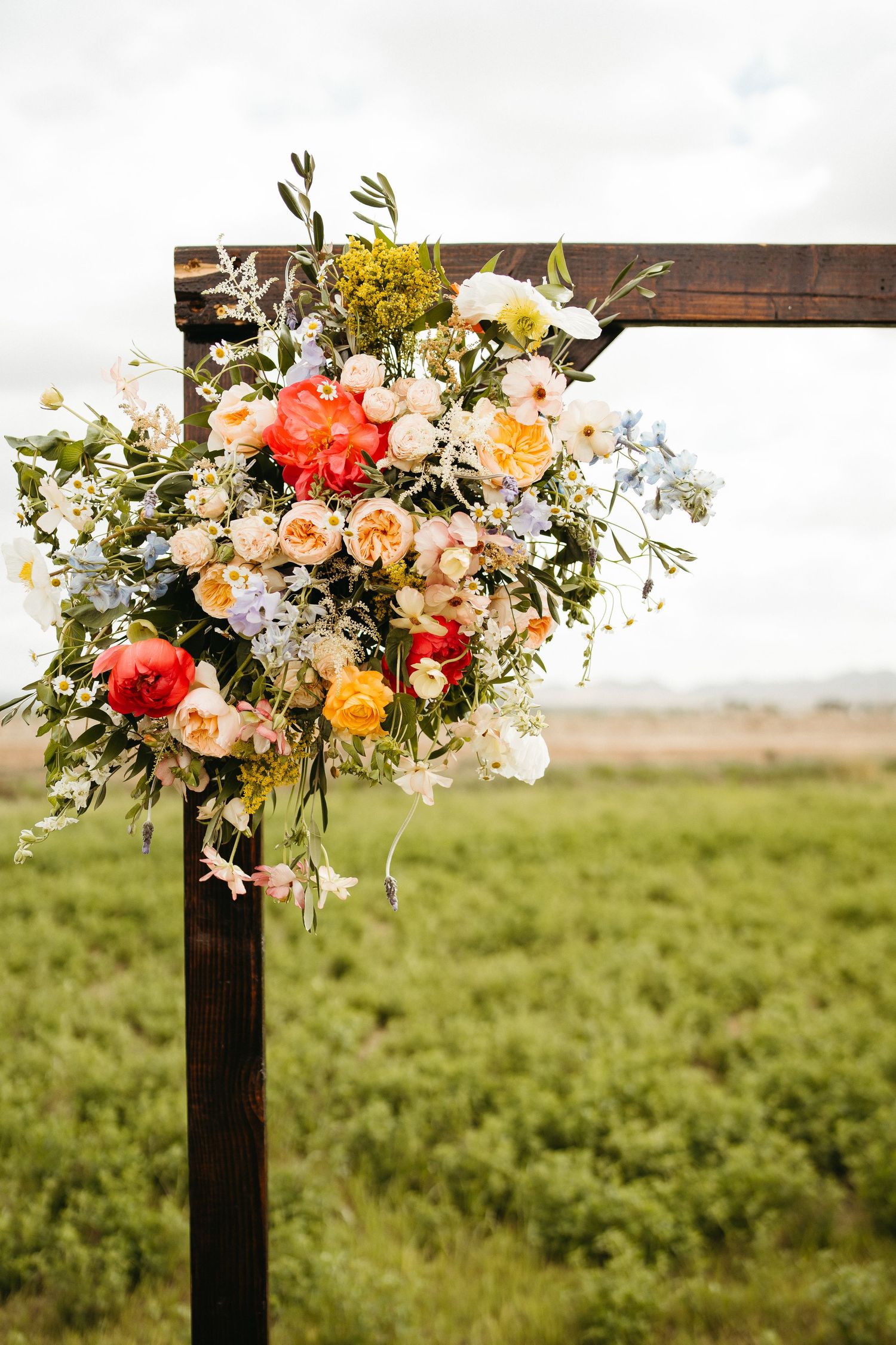 Colorful floral arrangement with roses and wildflowers adorns wooden wedding arch in outdoor setting.