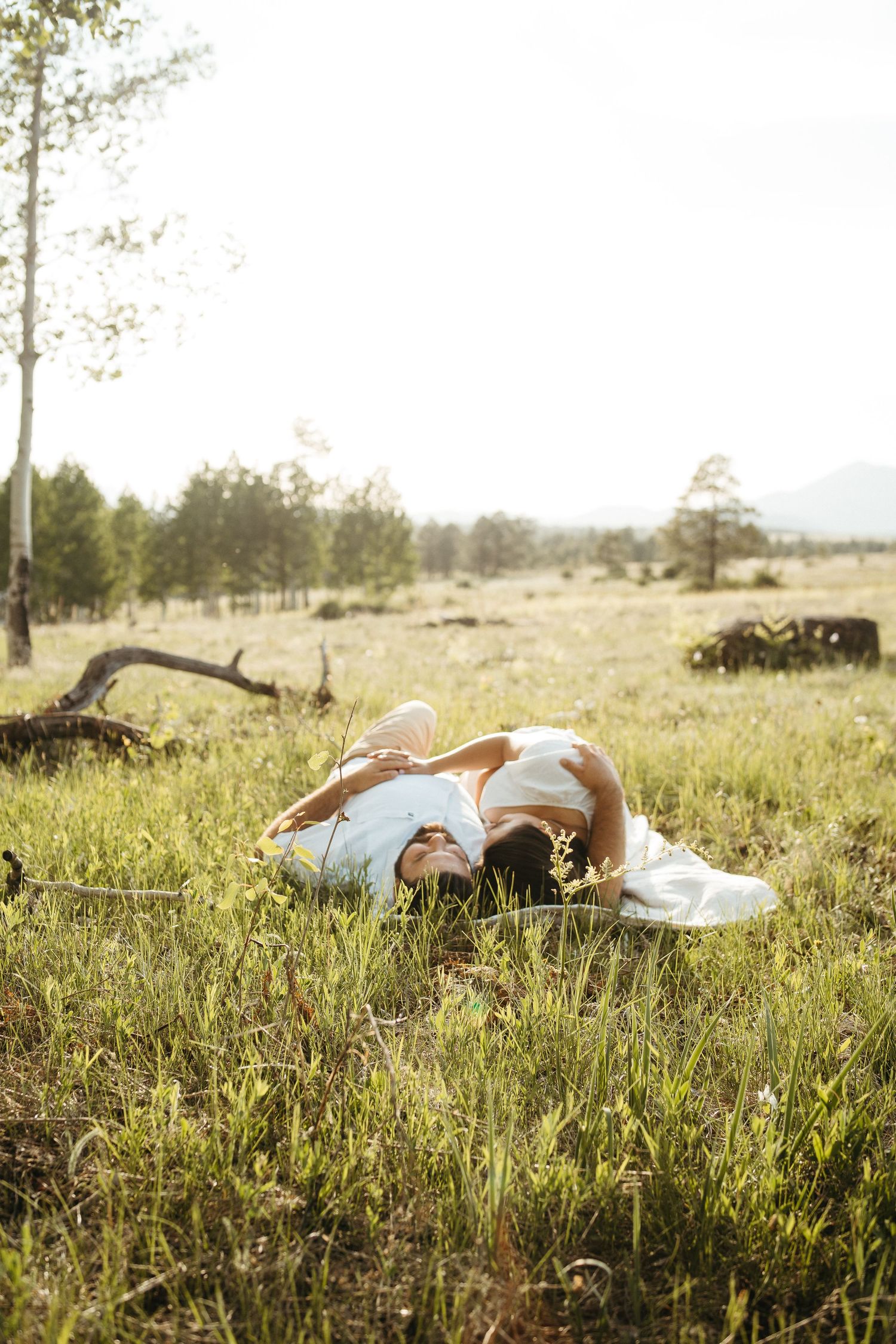 A couple lying together in a sunlit meadow surrounded by trees and mountains in the distance.