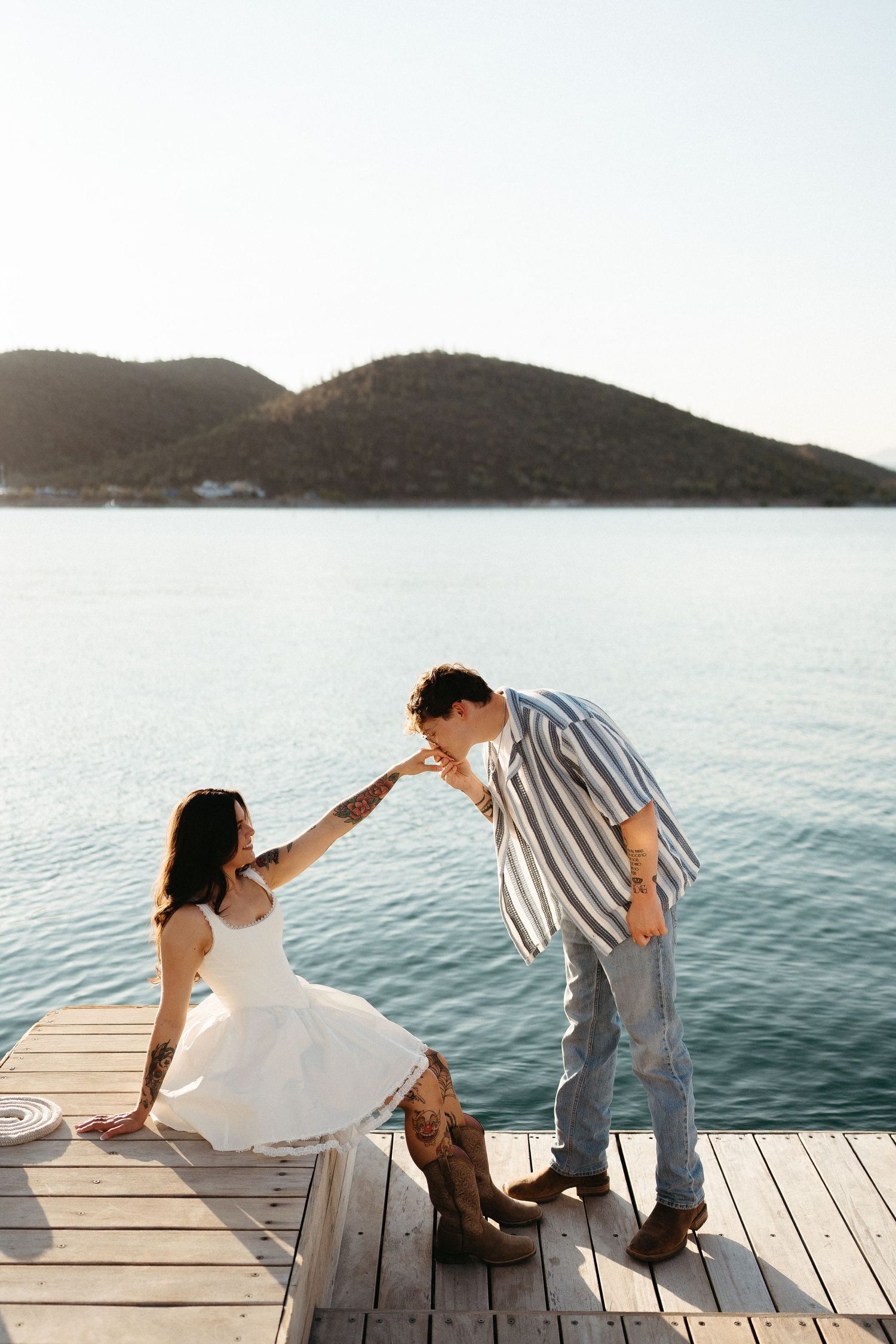 Two people dancing playfully together on a wooden dock by a calm lake with mountains in the background at sunset.