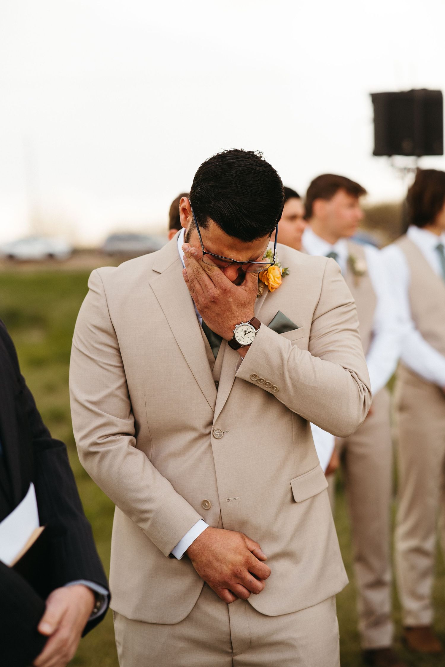 Emotional groom in beige suit wipes tears during outdoor wedding ceremony.