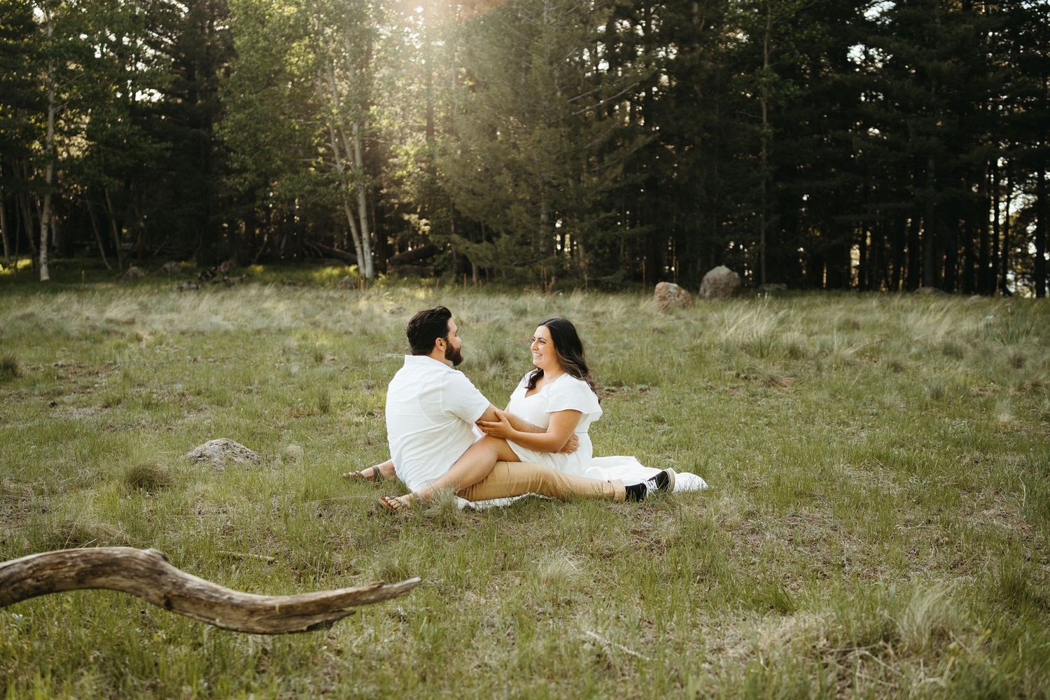Couple having an intimate moment while sitting in a sunlit meadow surrounded by evergreen forest.