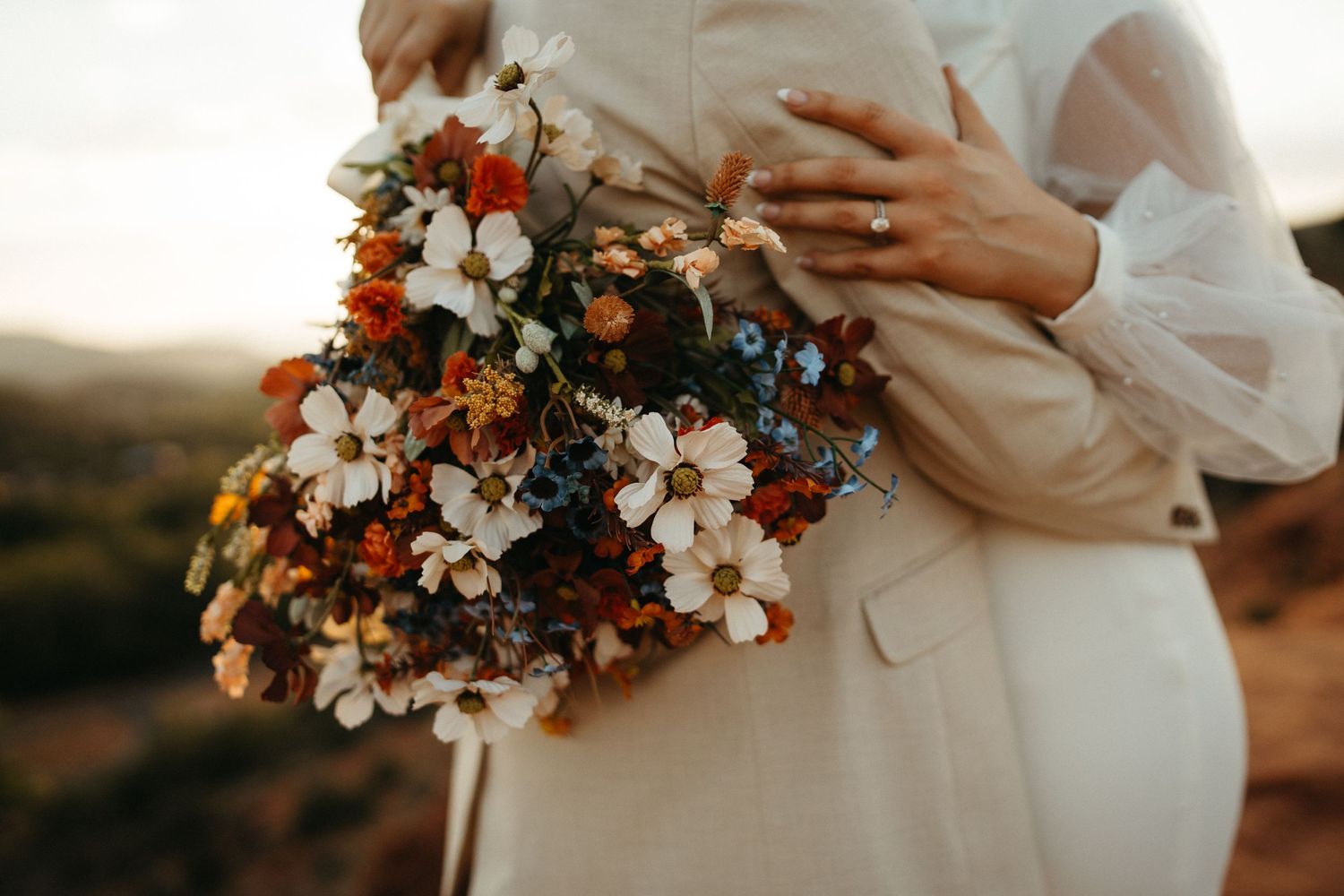 Bride holding rustic bouquet with orange and white flowers against sunset backdrop.