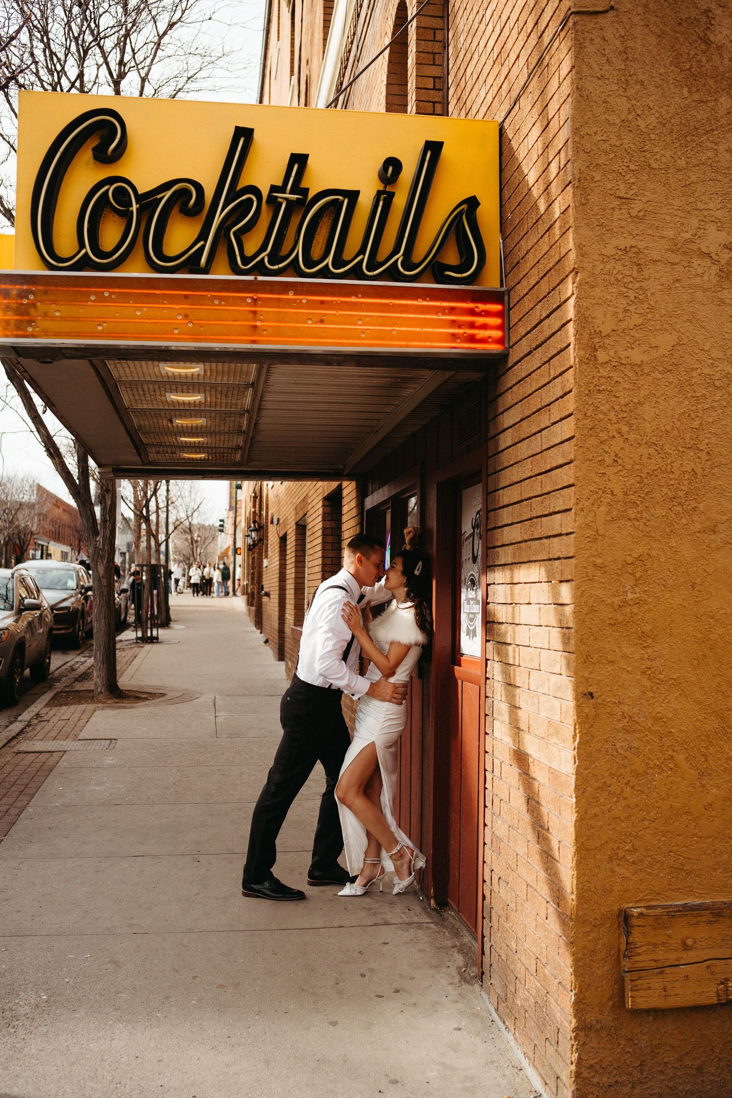 A romantic couple embraces beneath a vintage yellow cocktail sign on an urban sidewalk.