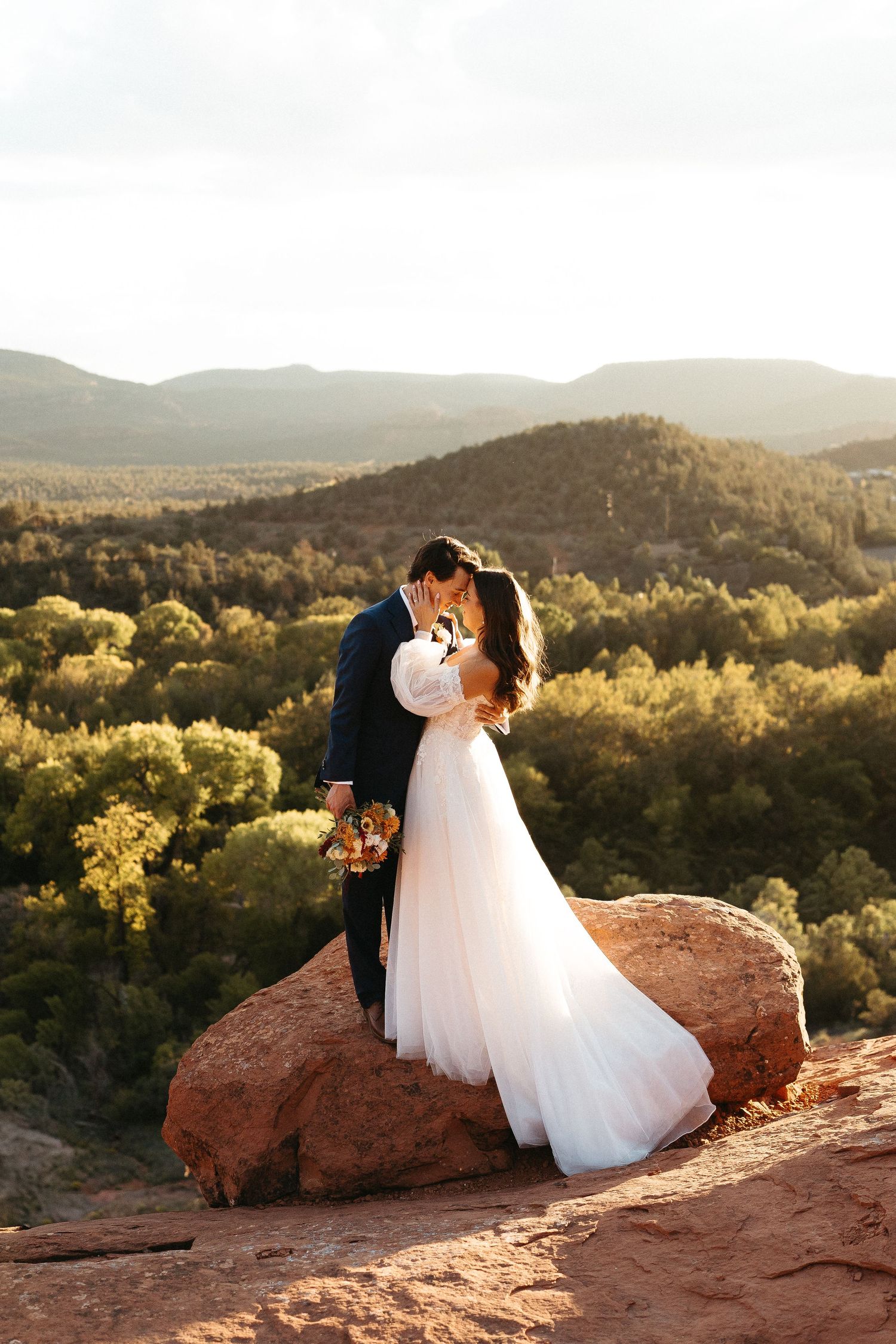 A newlywed couple embraces on a red rock overlook with golden sunlit mountains and desert landscape in the background.