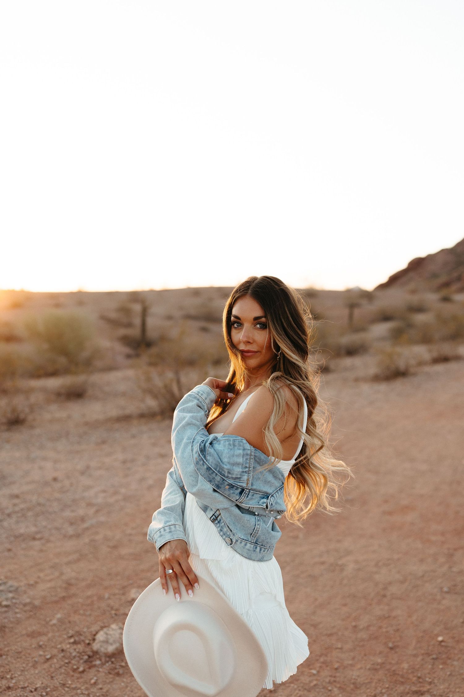 A person in a denim jacket and white dress poses in a desert landscape at sunset, holding a white hat.
