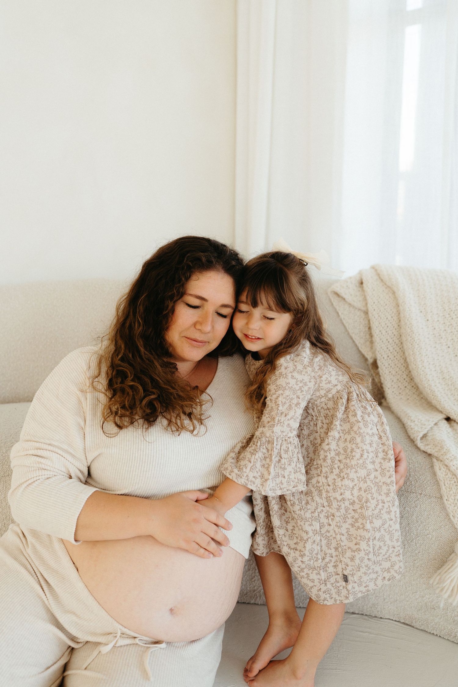 A pregnant woman in a white sweater shares tender moments with a young child in a floral dress on a light-colored couch.