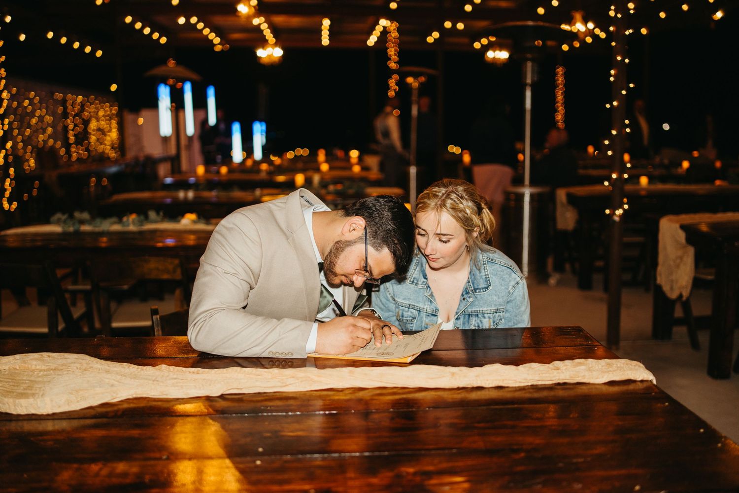 A couple shares an intimate moment while looking at a book together at a rustic wooden table in a warmly lit venue.