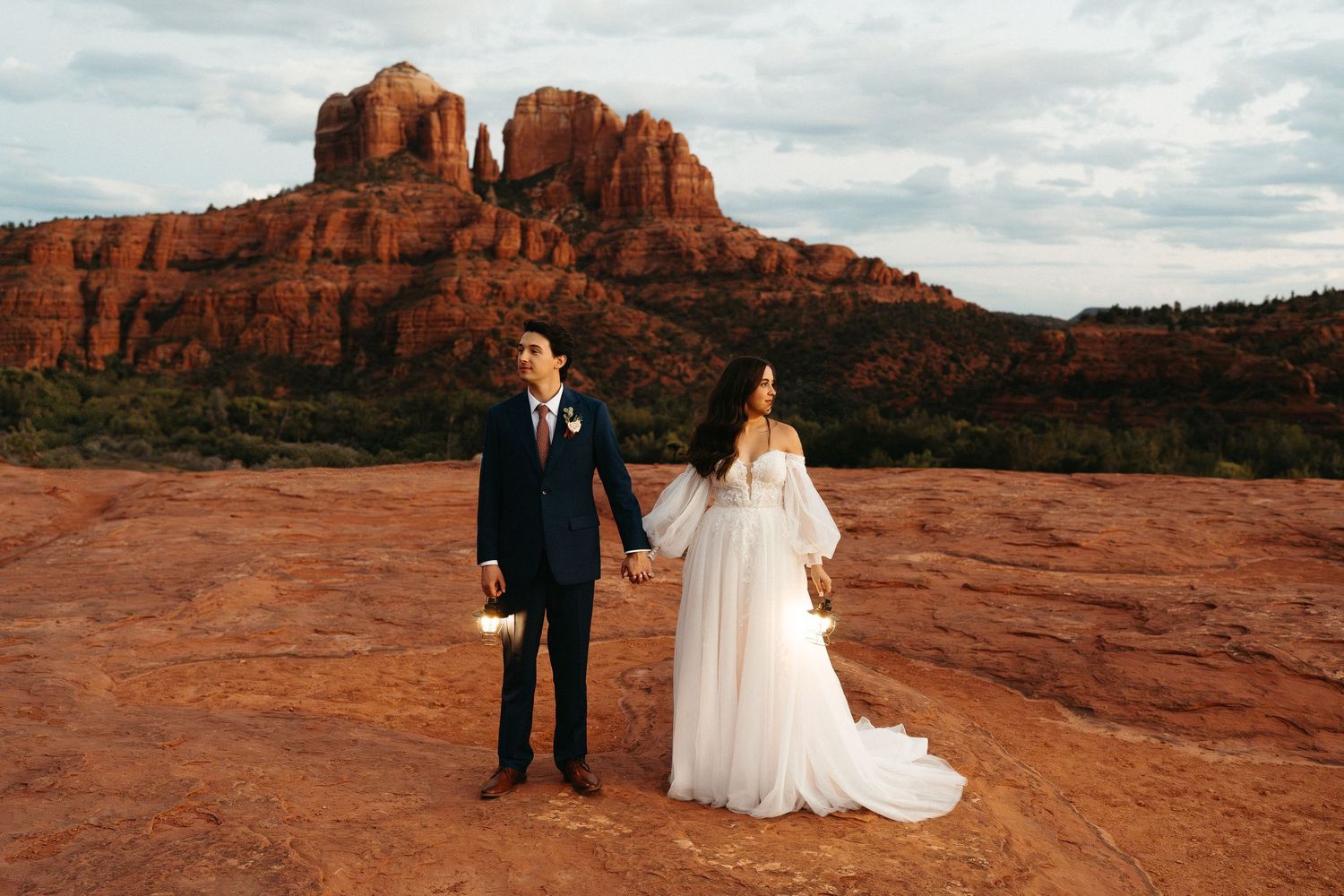 A couple in wedding attire stands on red rocks with majestic Cathedral Rock formations in Sedona, Arizona at sunset.