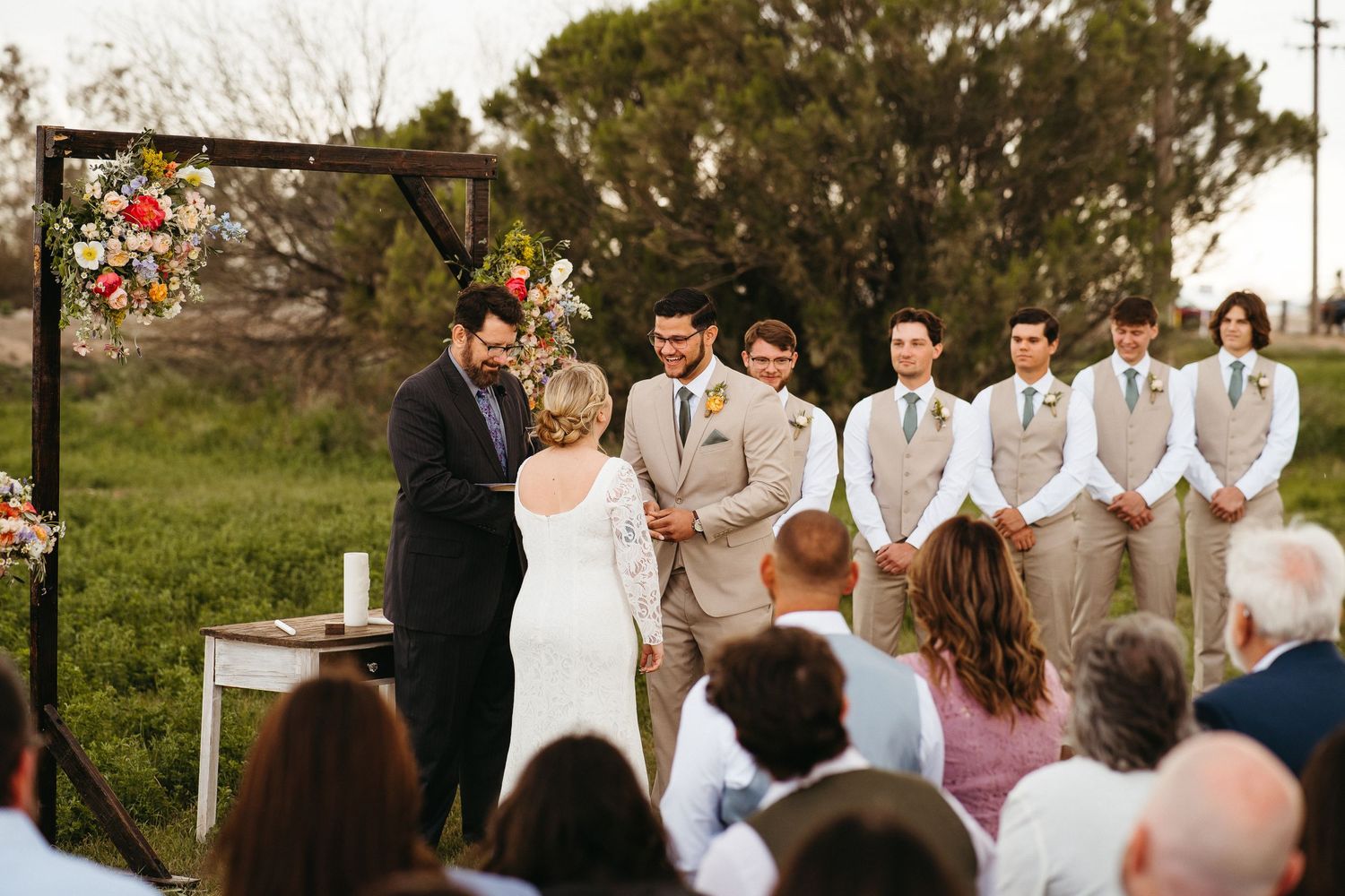 Outdoor wedding ceremony under wooden arch with floral arrangements and guests seated on lawn.