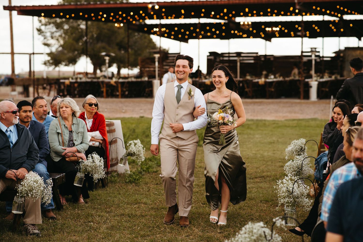 Wedding ceremony processional on grassy outdoor venue with string lights overhead.