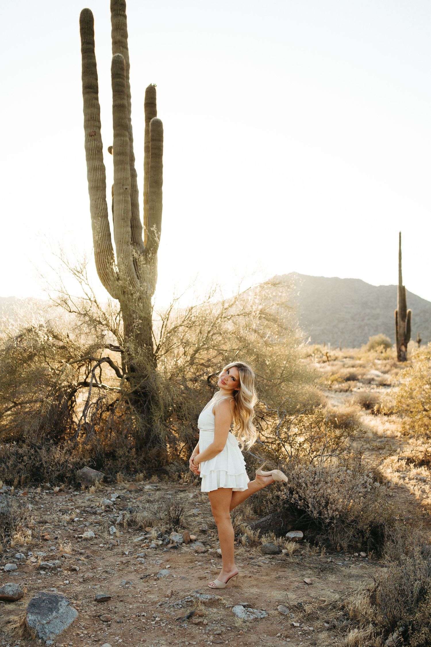 Person in white dress posing against desert landscape with towering saguaro cactus at sunset.