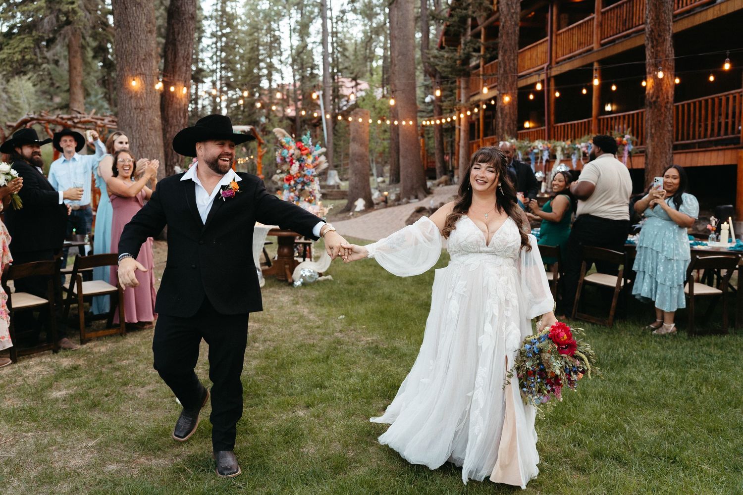 A joyful outdoor wedding celebration at a rustic venue with string lights and forest backdrop.