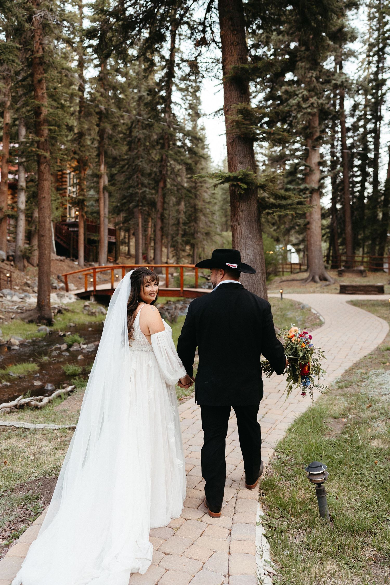 A couple walks along a winding stone path through a forest of tall pine trees at an outdoor wedding venue.