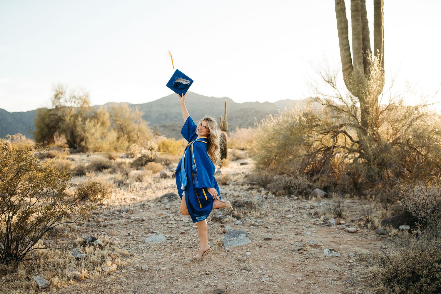 Graduate celebrating by tossing cap in desert landscape with towering saguaro cactus.